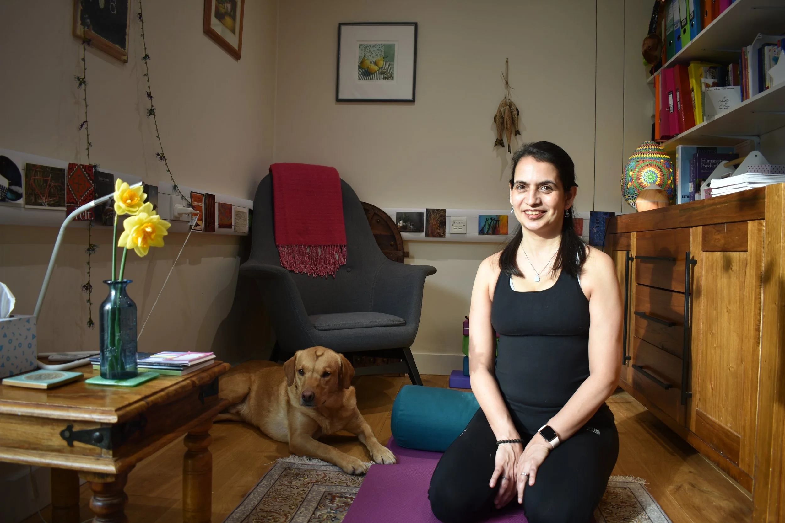 Sophia Armstrong kneeling on a purple yoga mat in a cosy, decorated room with a brown dog lying on a small rug, a armchair with a red blanket, a wooden sideboard, colorful books, and a lamp. The woman is smiling at the camera.