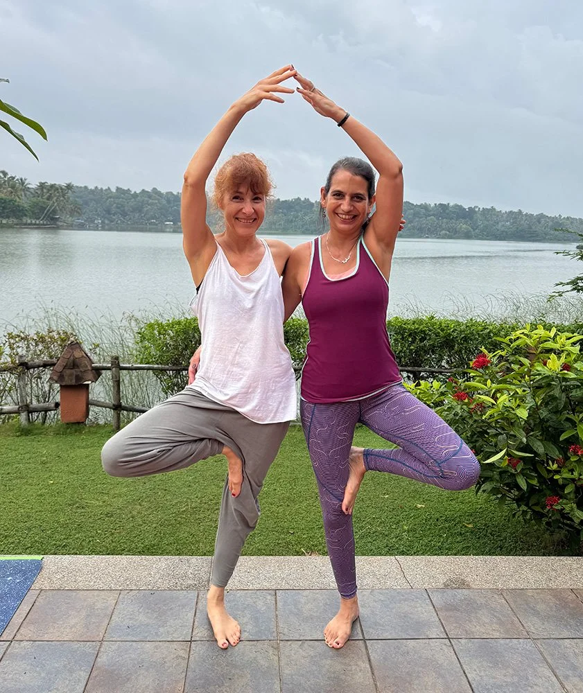 Two women practicing yoga outdoors by a lake, balancing on one leg with hands together overhead, surrounded by greenery and a cloudy sky.