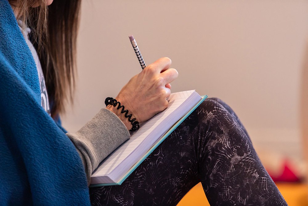 A person sitting and writing in a notebook with a pen, wearing a blue jacket and patterned leggings.