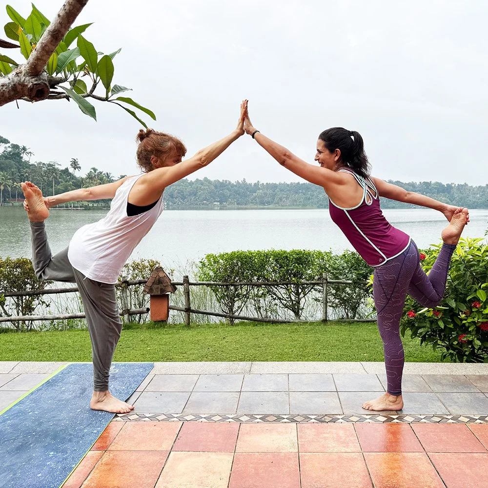 Two women practicing yoga outdoors near a lake, balancing in a pose with one leg lifted and hands together in a high-five position.