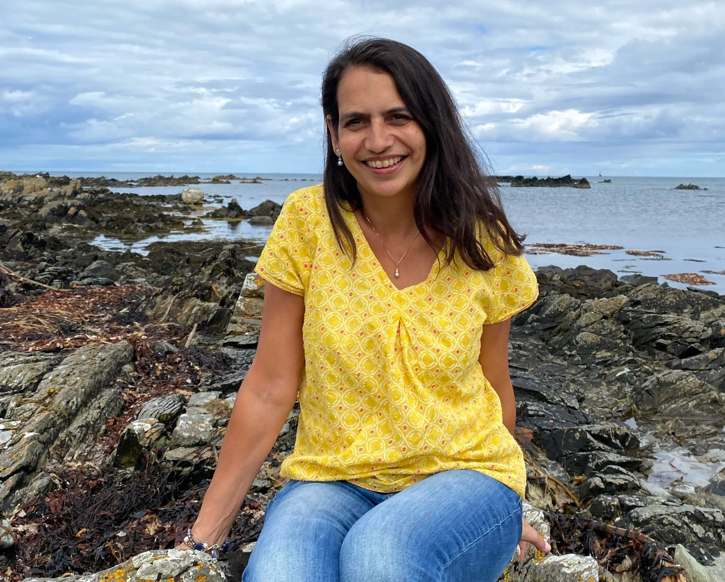 Sophia Armstrong sitting on rocky beach with ocean and cloudy sky in the background.