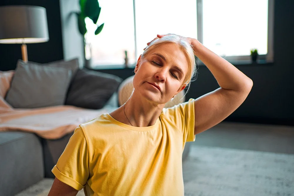 A woman in a yellow shirt is doing a yoga stretch in a cosy living room with a bed, pillows, and large windows.
