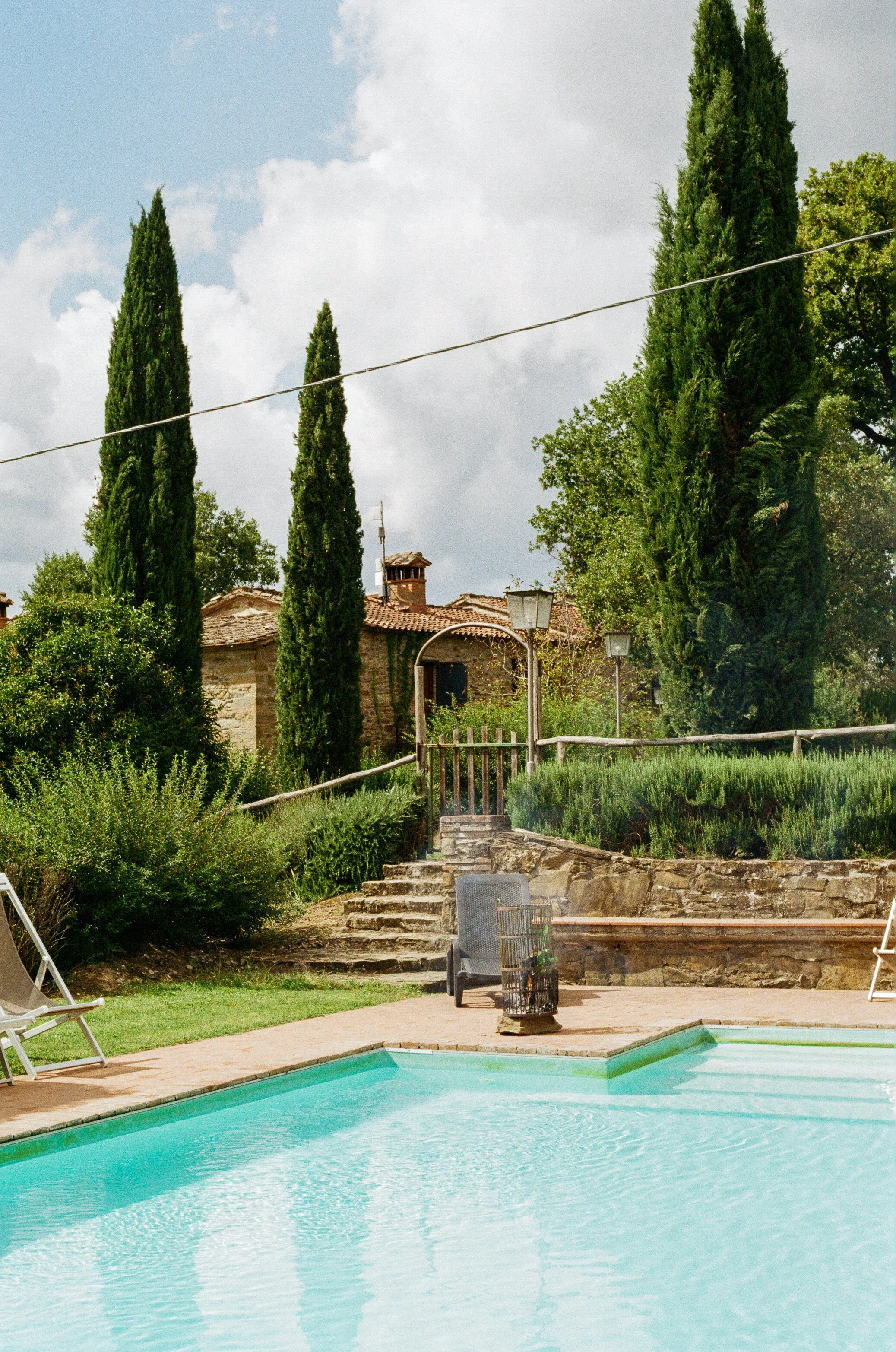 Outdoor swimming pool with lounge chairs, surrounded by greenery and cypress trees.