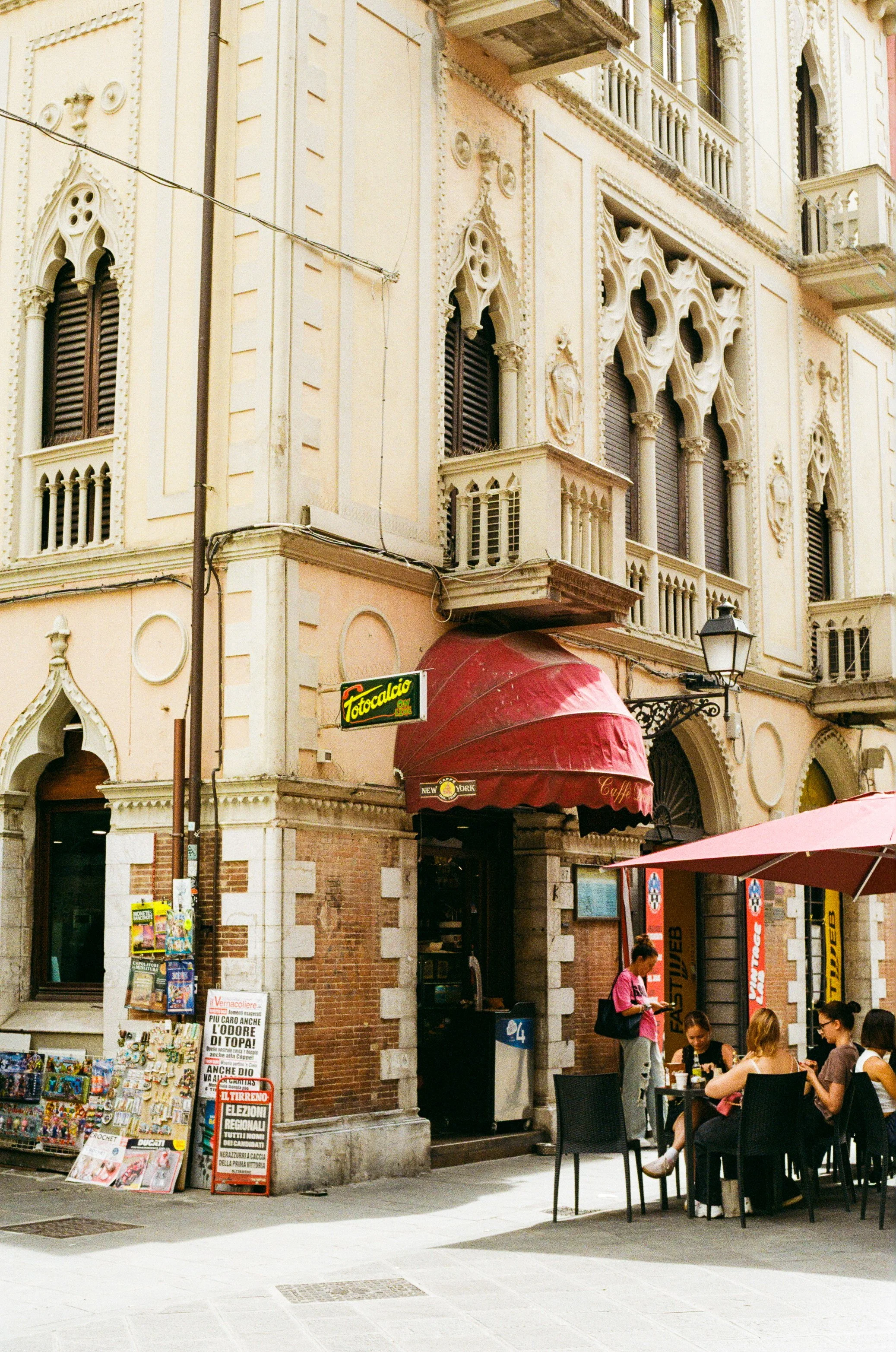 People sitting at an outdoor café in front of an ornate, historic building with arched windows and detailed stonework in a European city.
