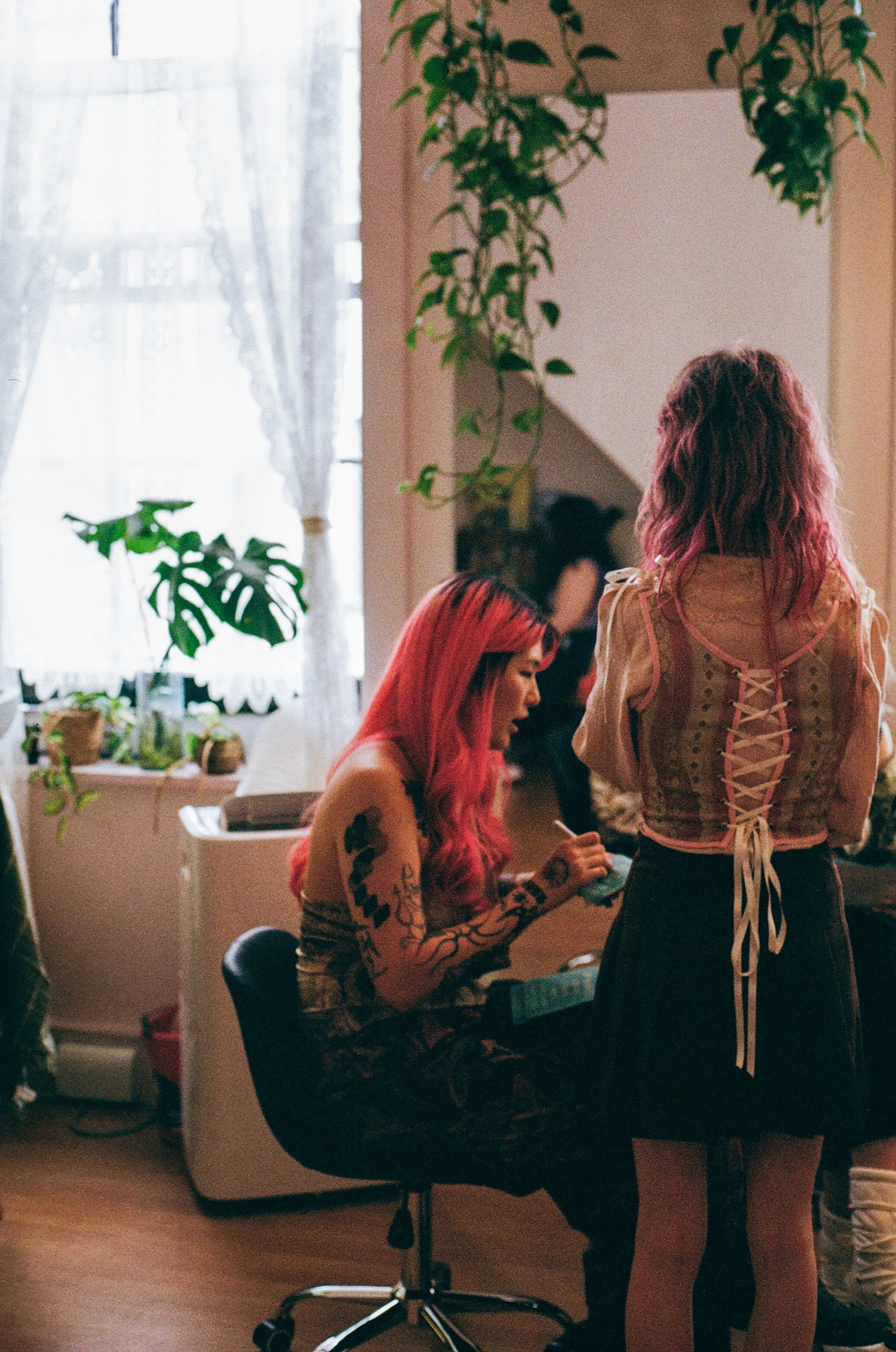 Two women with pink hair in a room with plants, one sitting at a desk writing and the other standing with her back to the camera, talking.