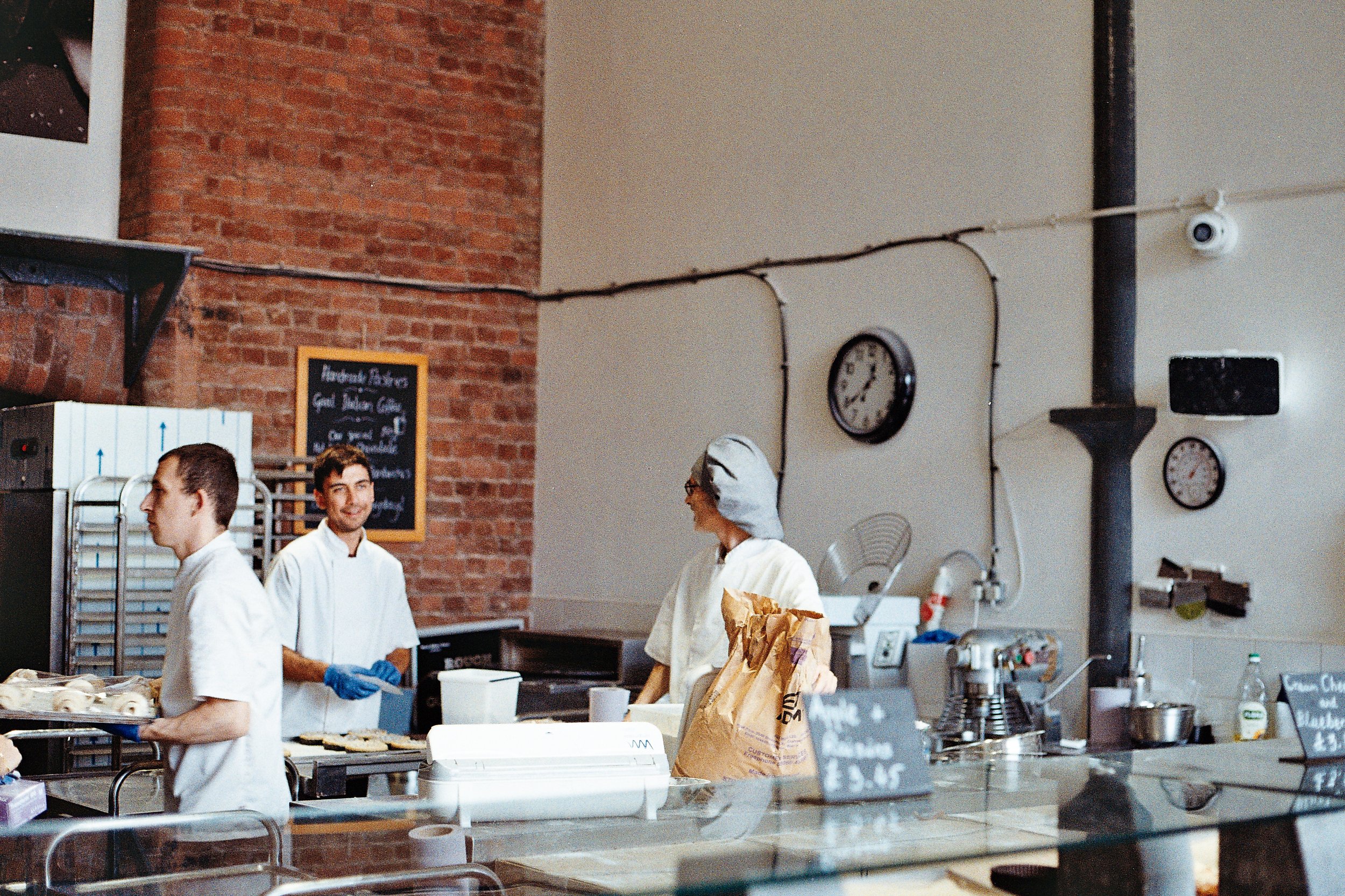 Three chefs working behind the counter in a restaurant kitchen, with brick and white walls, clocks, and kitchen equipment visible.