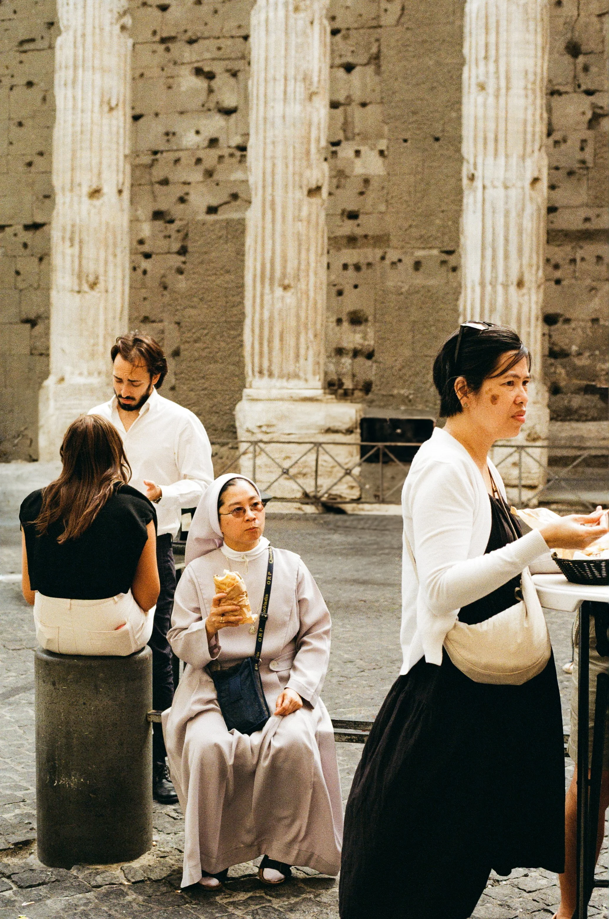 People sitting and standing in front of old stone columns at an outdoor location, with one woman eating and another woman looking at her phone.