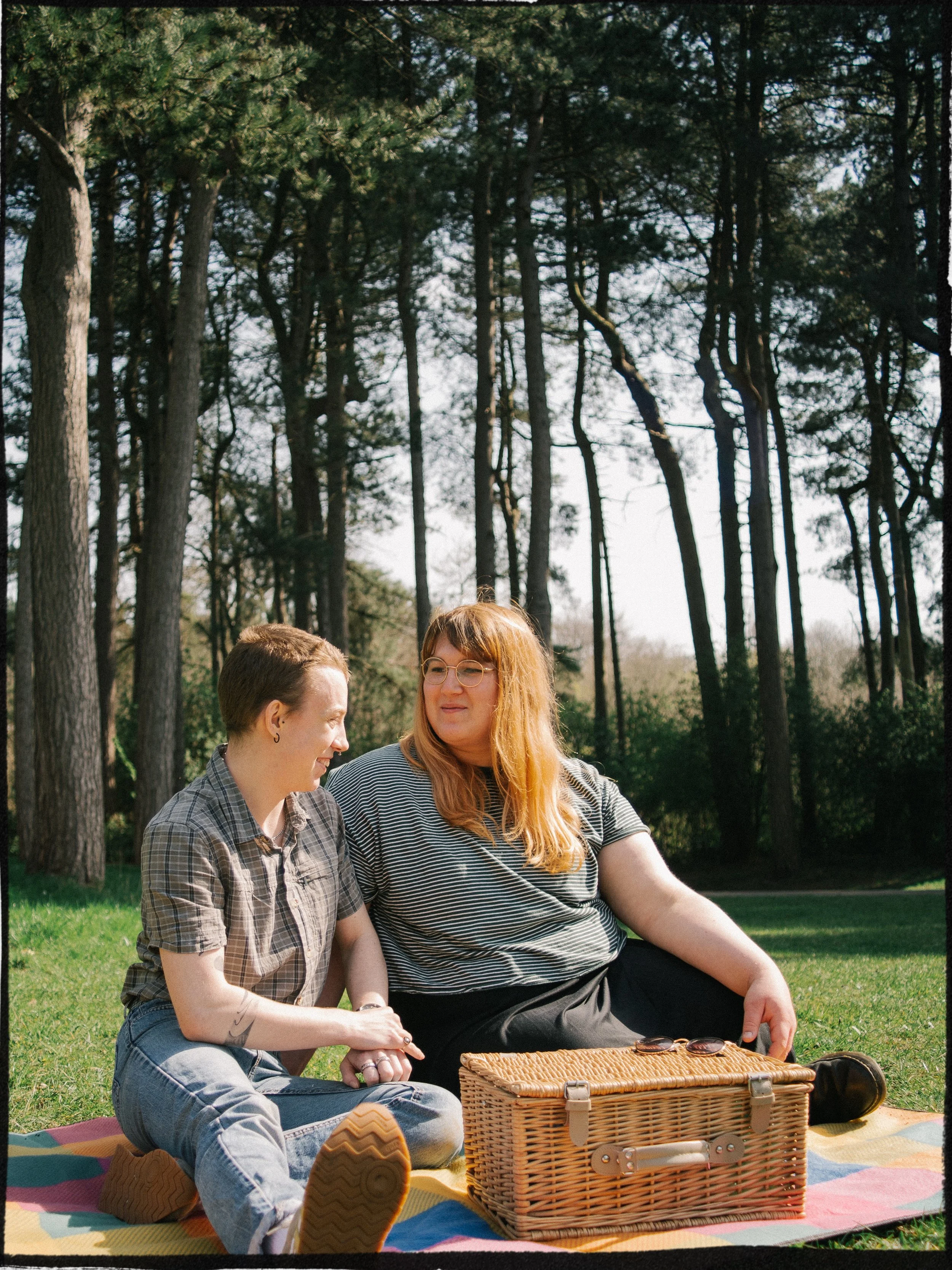 Two women sitting on a multicolored blanket outdoors with a wicker picnic basket, sunglasses, and other items. They are surrounded by tall trees and appear to be enjoying a sunny day.
