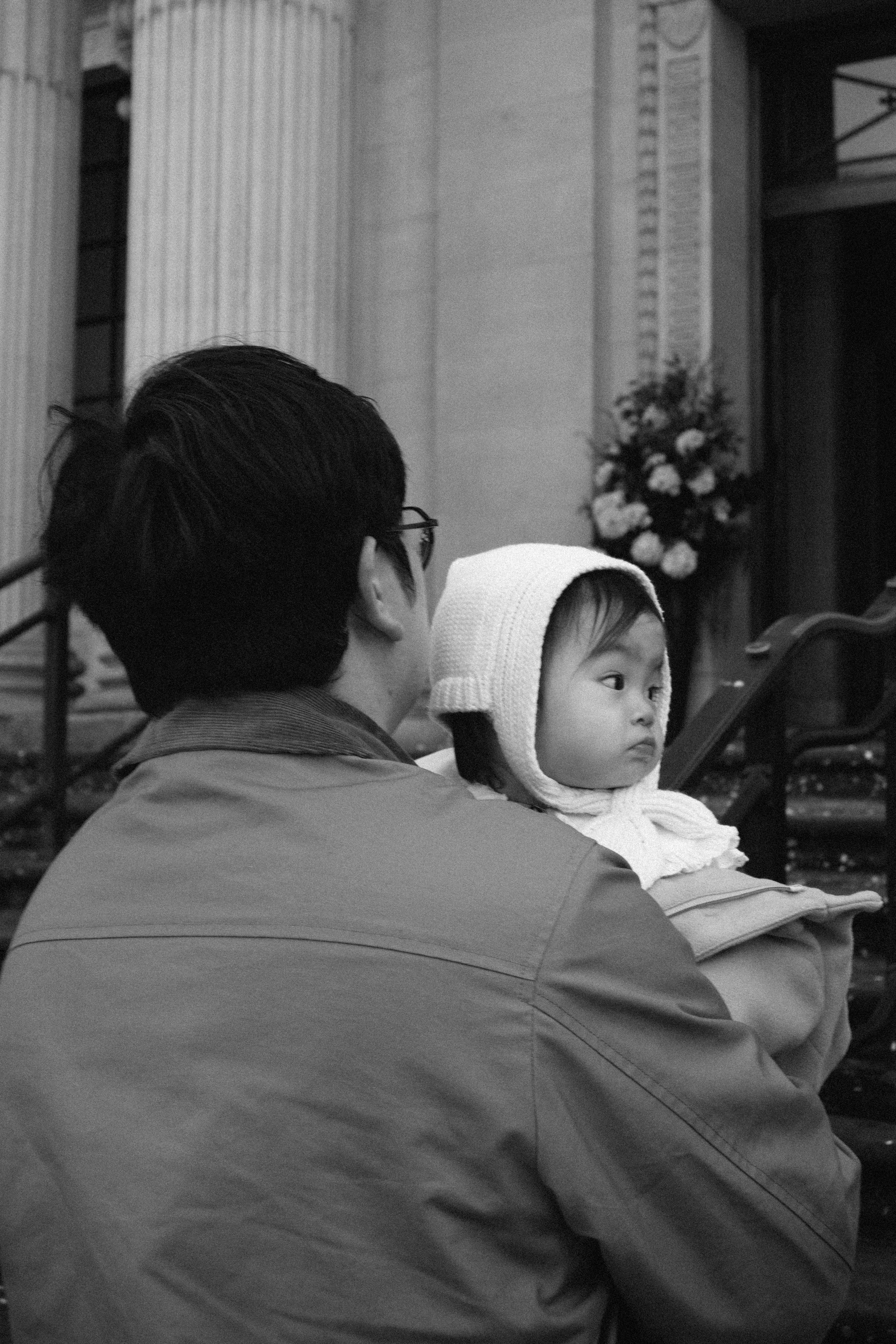 A man holding a young child, both facing sideways, in an indoor setting with floral arrangements and stairs in the background.