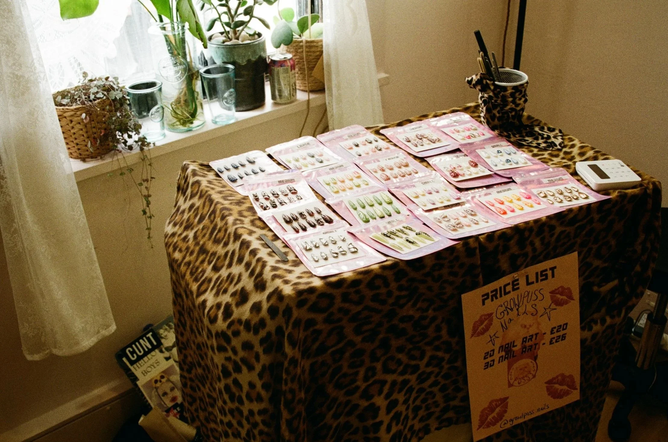 A display table covered with leopard print fabric holding various sets of jewelry and nail art designs, with a price list sign attached. The table is near a window with potted plants on the windowsill, and a cup with pens or makeup brushes on top.