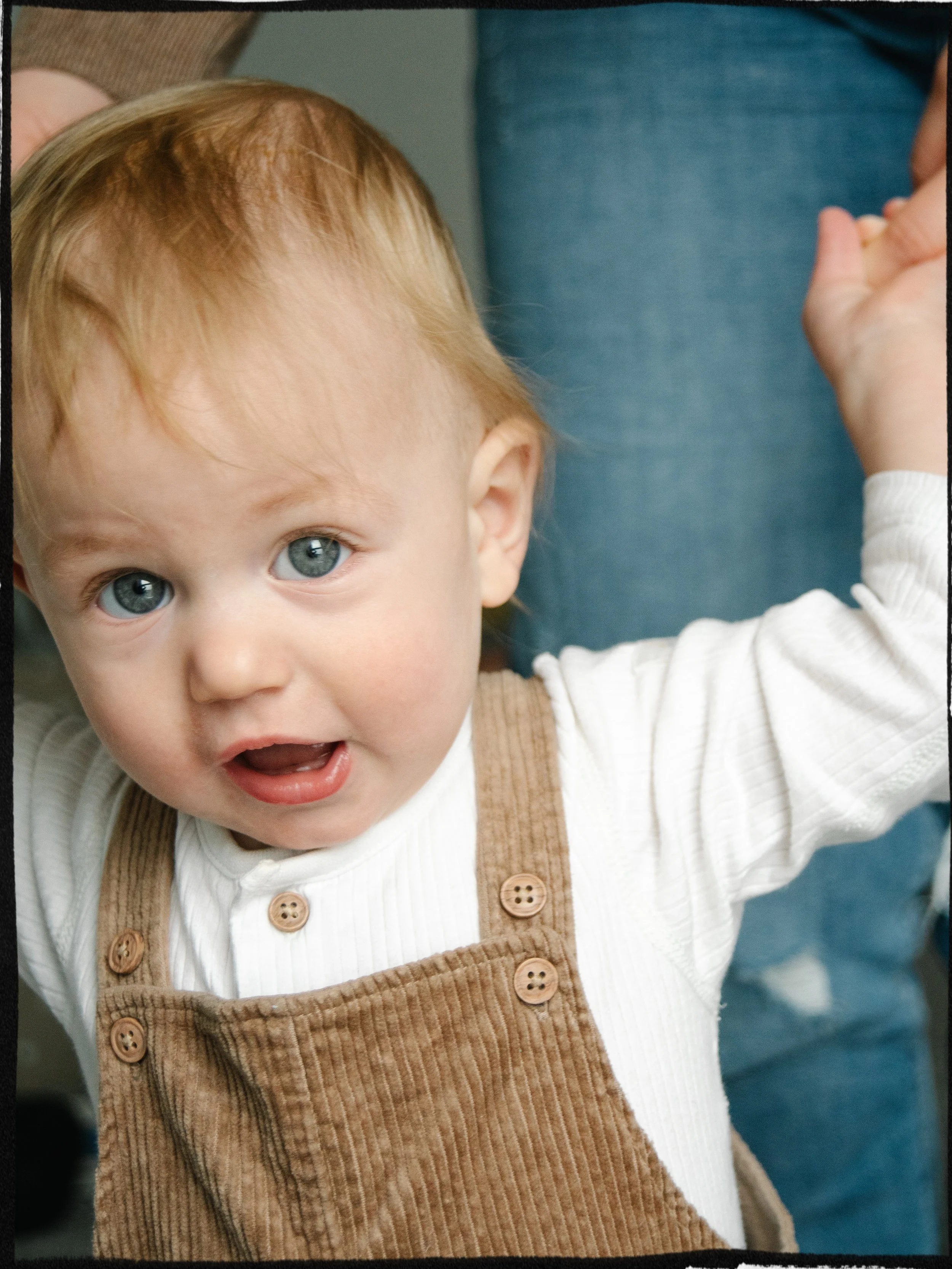 A young child with red hair and blue eyes, wearing a white long-sleeve shirt and brown corduroy overalls, looks at the camera with a curious expression and mouth slightly open.