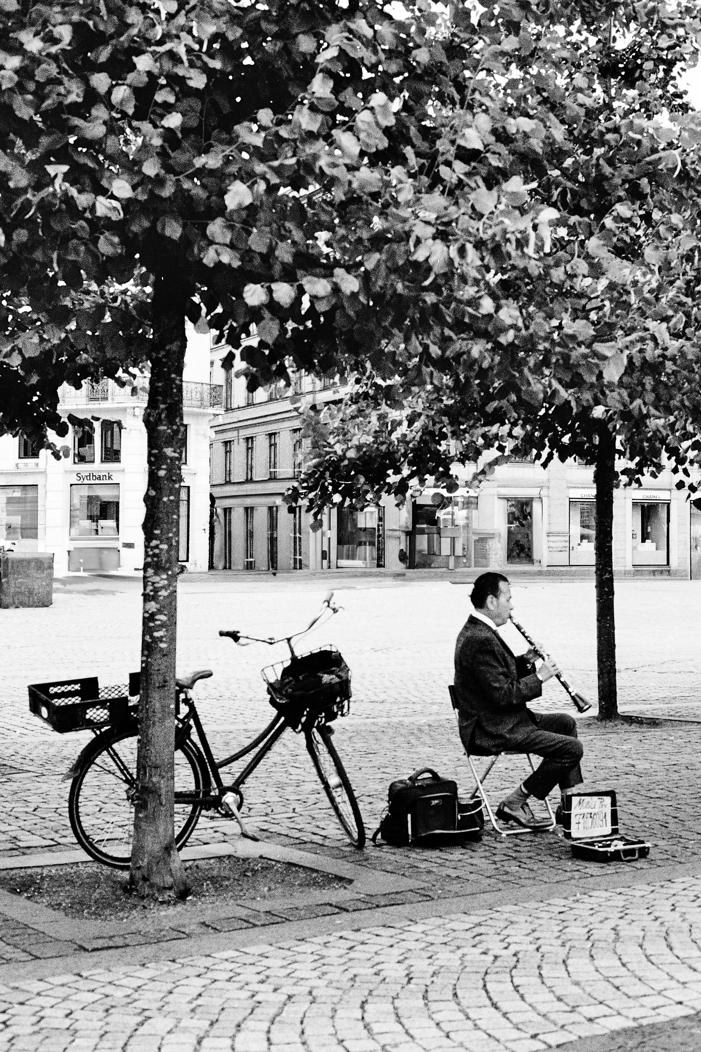 A man sitting on a folding chair playing a clarinet on a cobblestone street, with an open case for tips in front of him, and a bicycle parked against a tree nearby. There are trees, buildings, and shopfronts in the background.