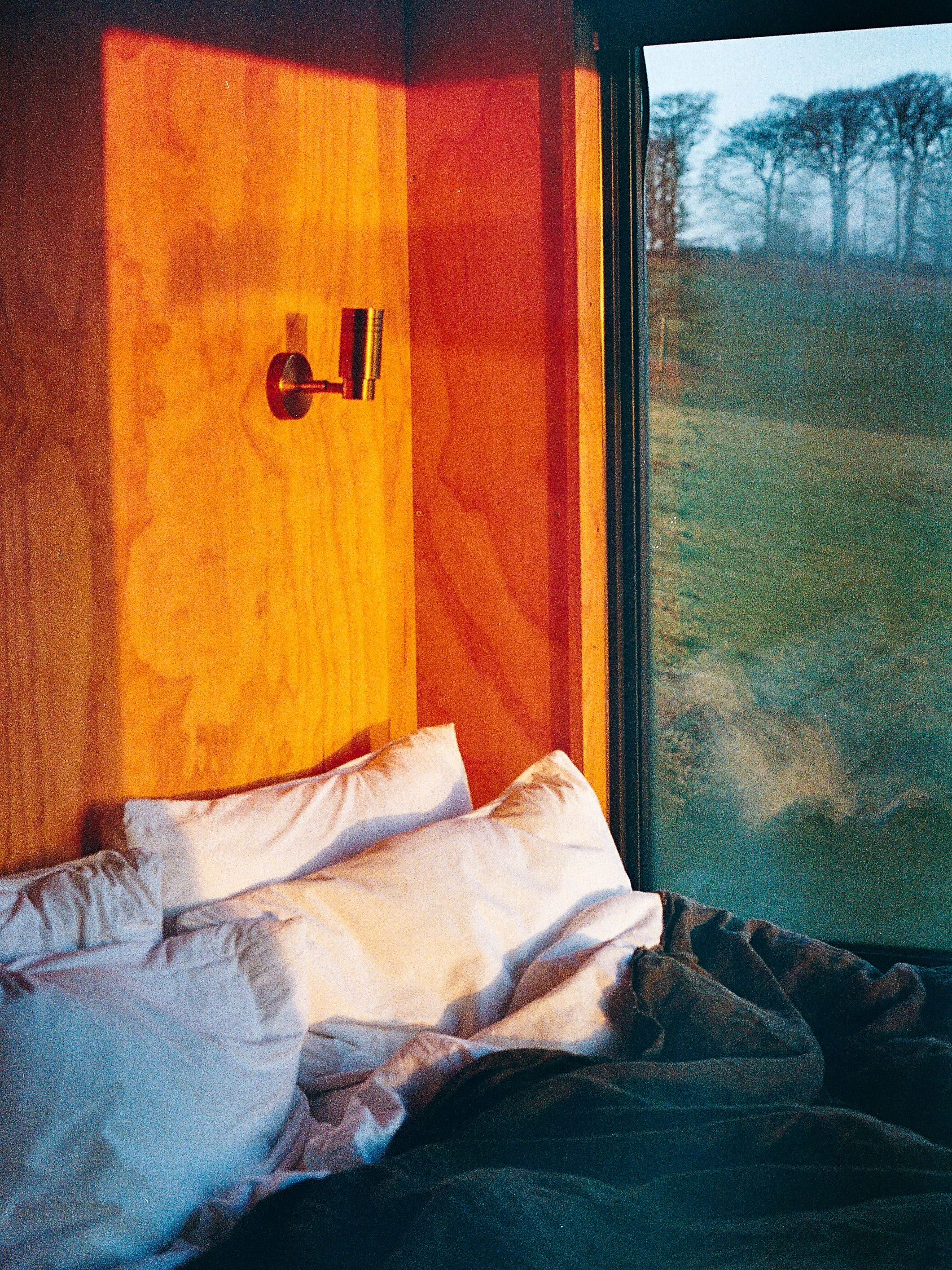 A cozy bedroom with white pillows and a blanket on a bed, a wooden headboard, a window showing a landscape with trees, and a small wall-mounted light fixture.