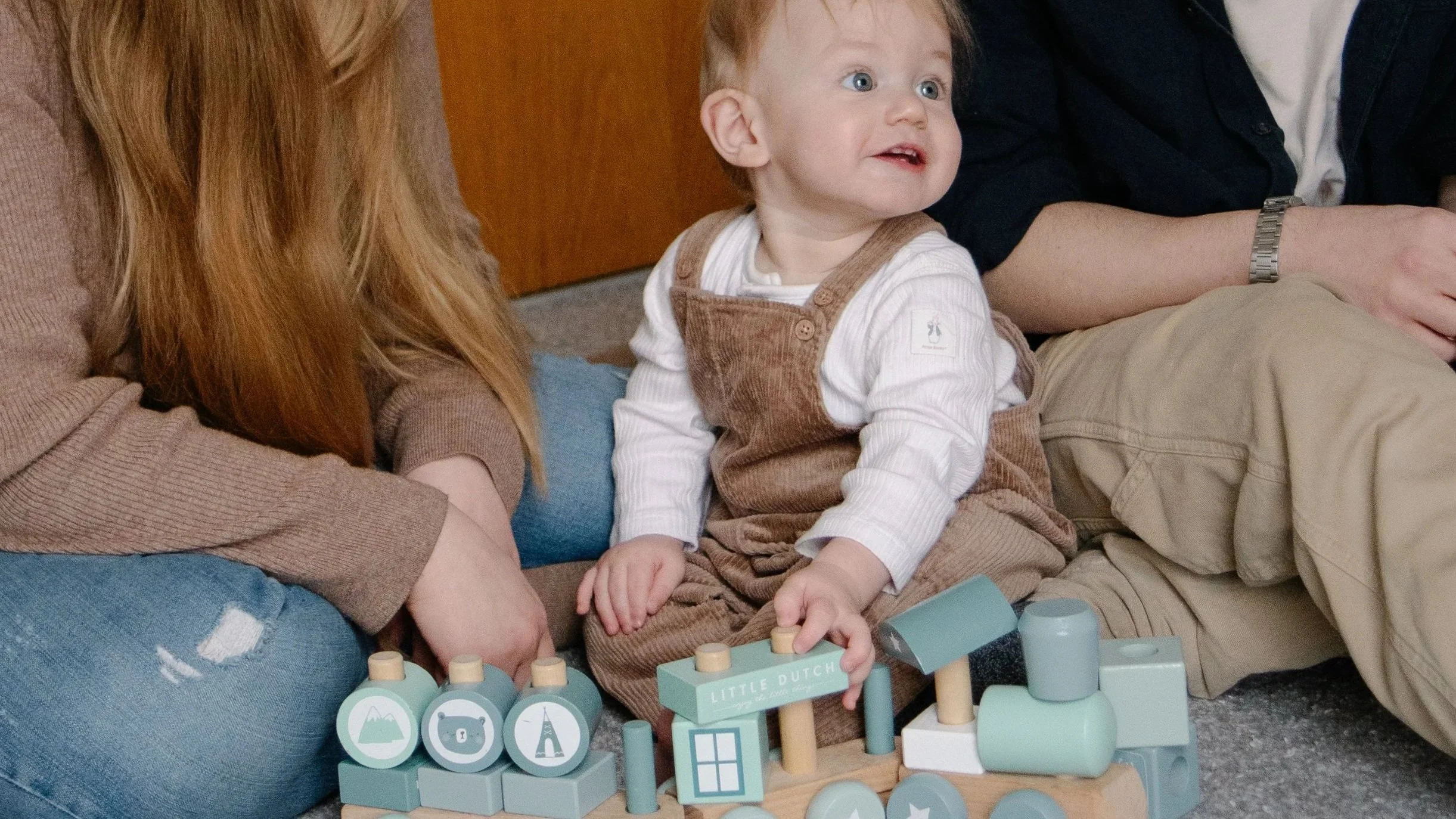 Young child with light brown hair, blue eyes, wearing a white long sleeve shirt and brown overalls, sitting on the floor with toys and two adults, one with long red hair and the other with a watch and beige pants, nearby.
