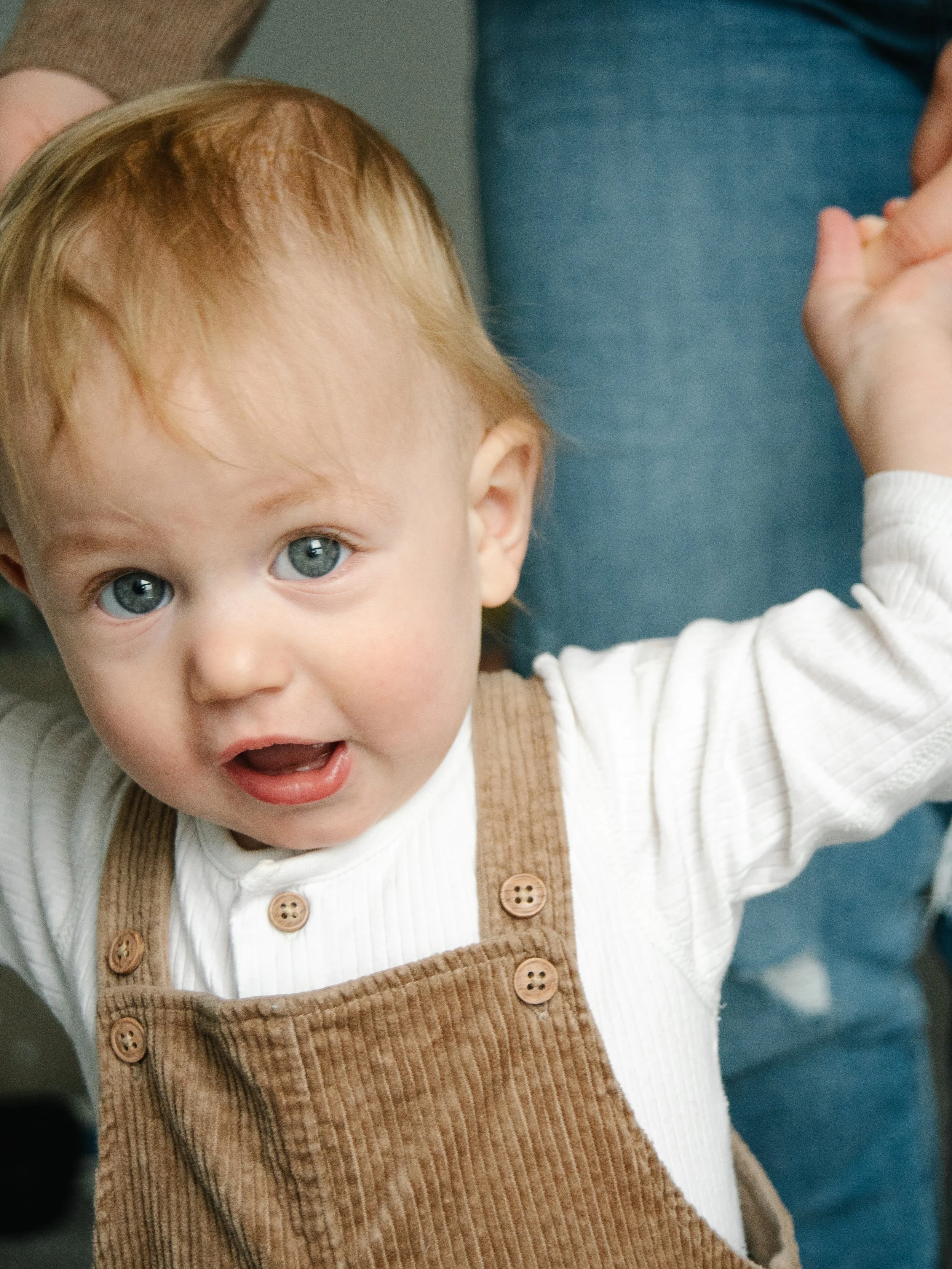 A young child with light skin, blond hair, and blue eyes raising their arm, wearing a white long-sleeve shirt and tan corduroy overalls, with an adult partially visible in the background.