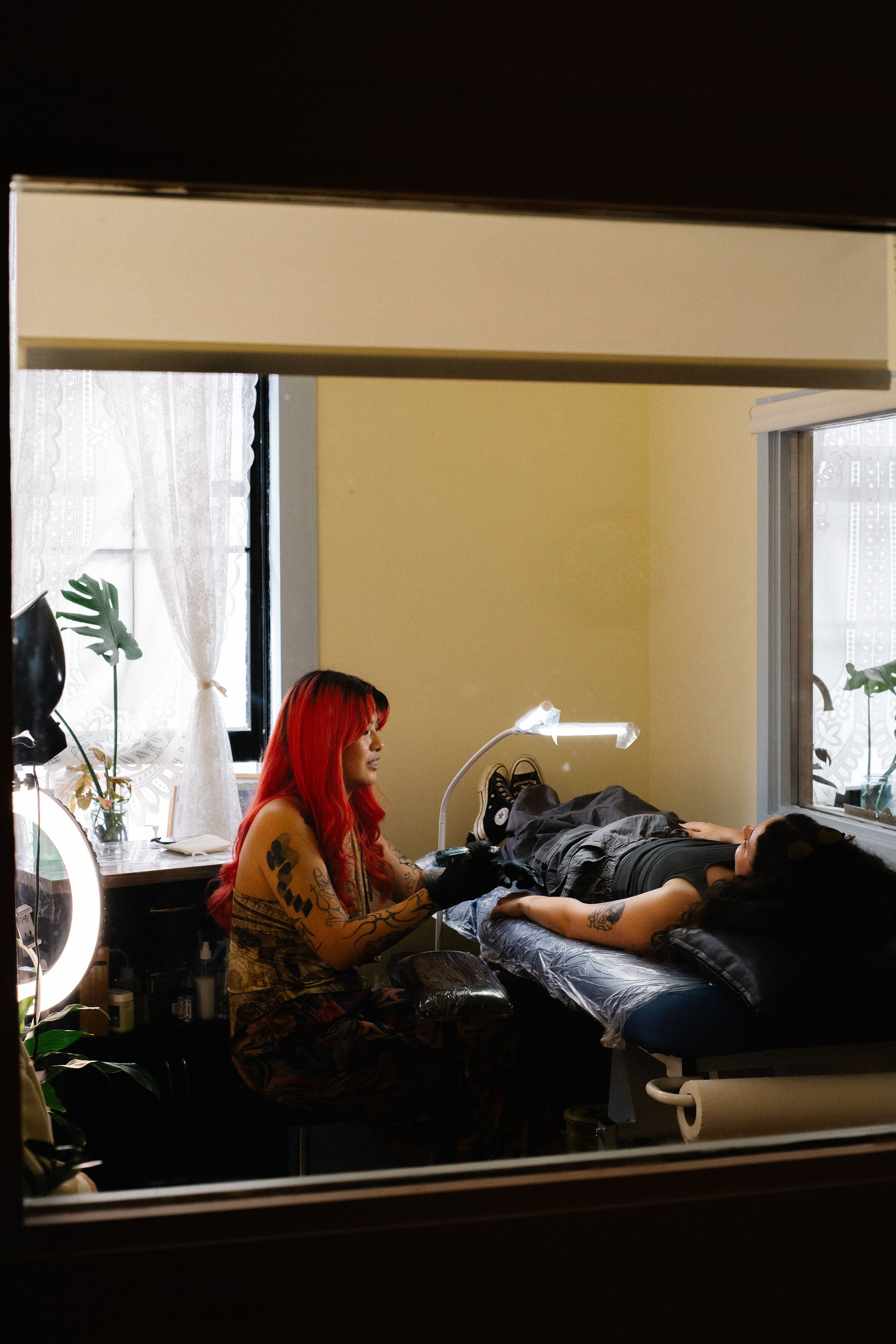 Tattooed woman with red hair giving a tattoo to a woman lying on a tattoo chair, inside a tattoo studio
