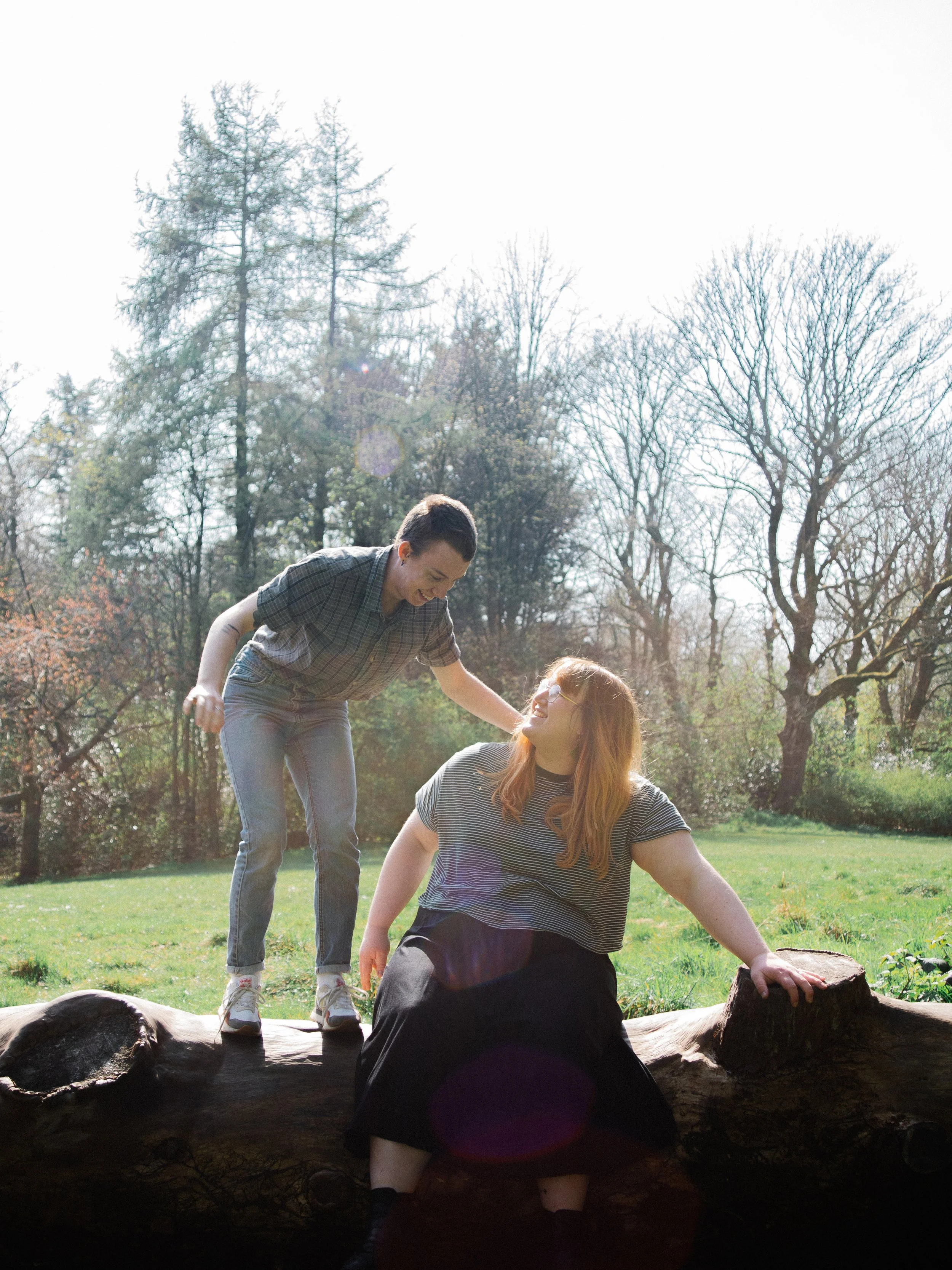 A man and a woman outdoors in a park on a sunny day, the man is standing on a fallen log leaning toward the woman, who is sitting on the log, both smiling and engaging with each other, with trees and grass in the background.
