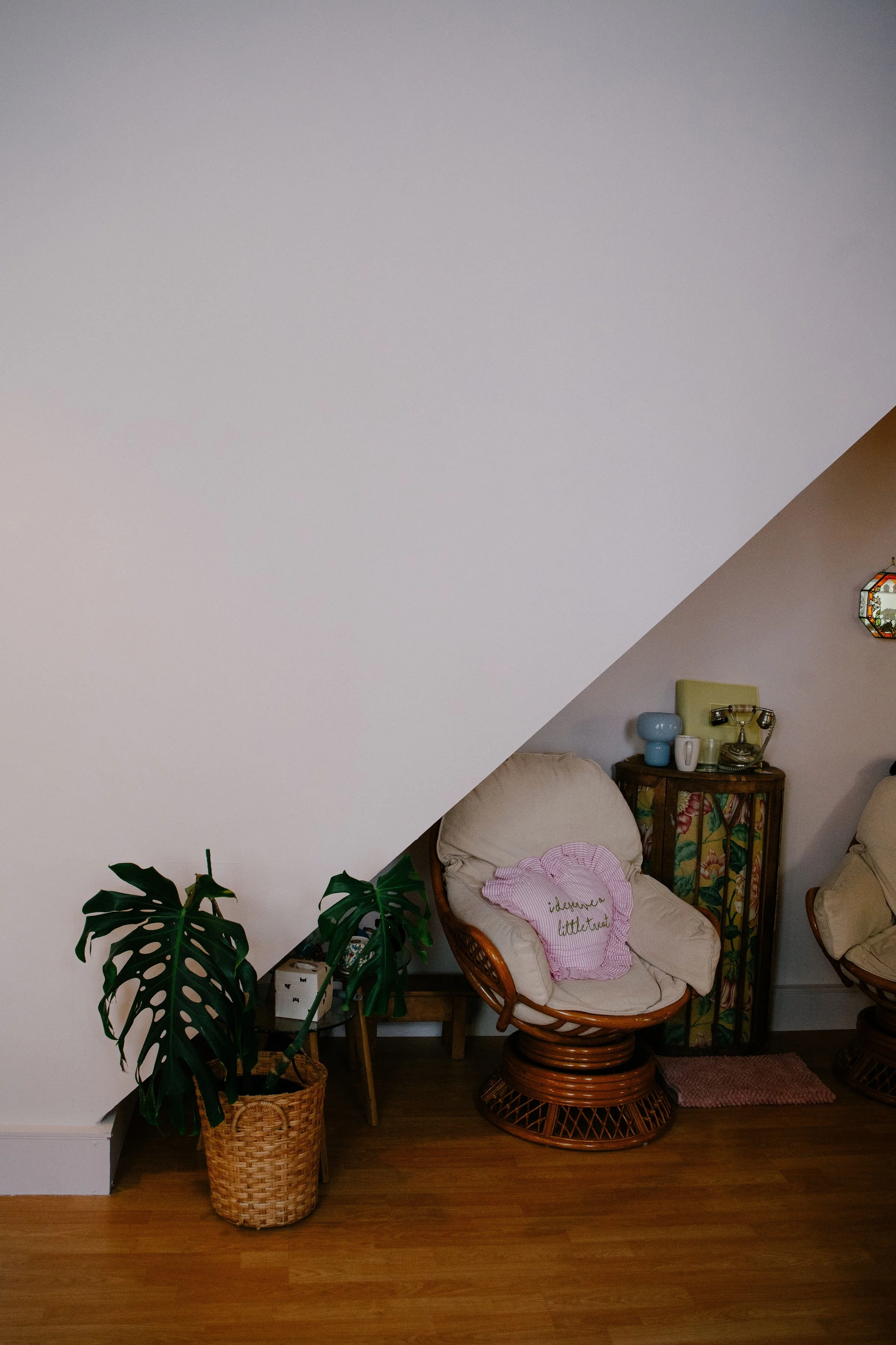 Living room corner with a rattan chair, a pink heart-shaped pillow, a tall floral cabinet, and a potted Monstera plant in a wicker basket, under a sloped ceiling.