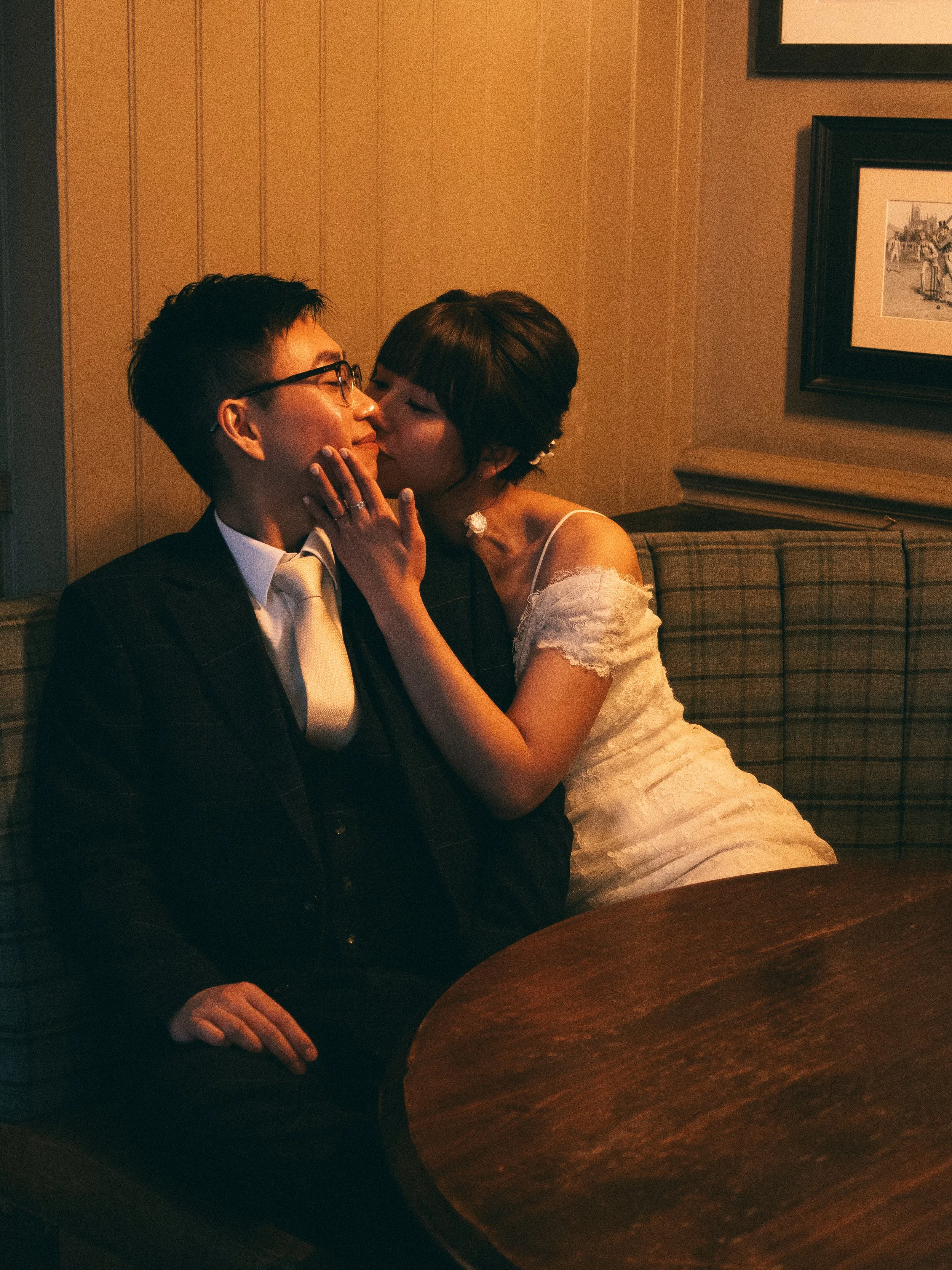 A couple sharing a romantic moment in a cozy, dimly lit room. The woman is leaning in to kiss the man's cheek while holding his face and touching his chin. The man is sitting on a checked sofa, wearing glasses and a suit, smiling softly. There are framed pictures on the wall behind them.