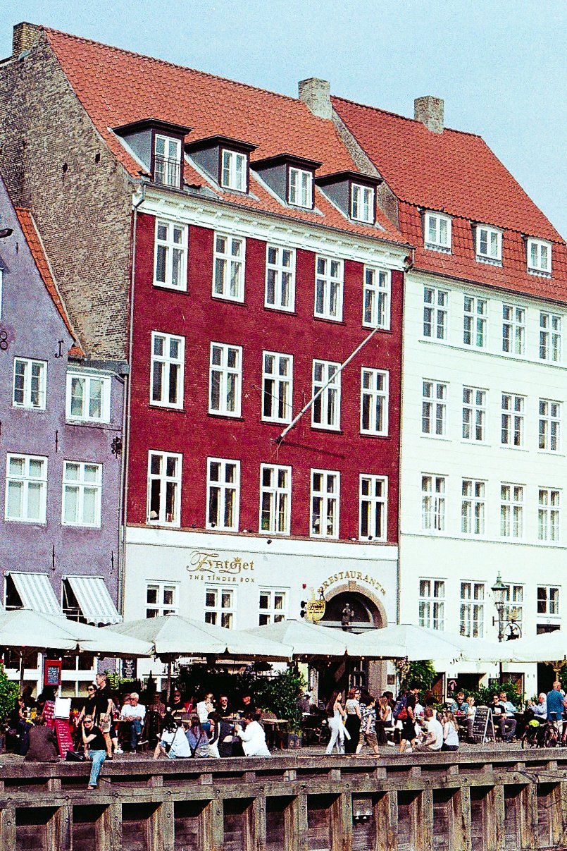 Street scene with outdoor seating outside a restaurant in front of colorful multi-story buildings with red, purple, and white facades, and a crowd of people walking and sitting under umbrellas.