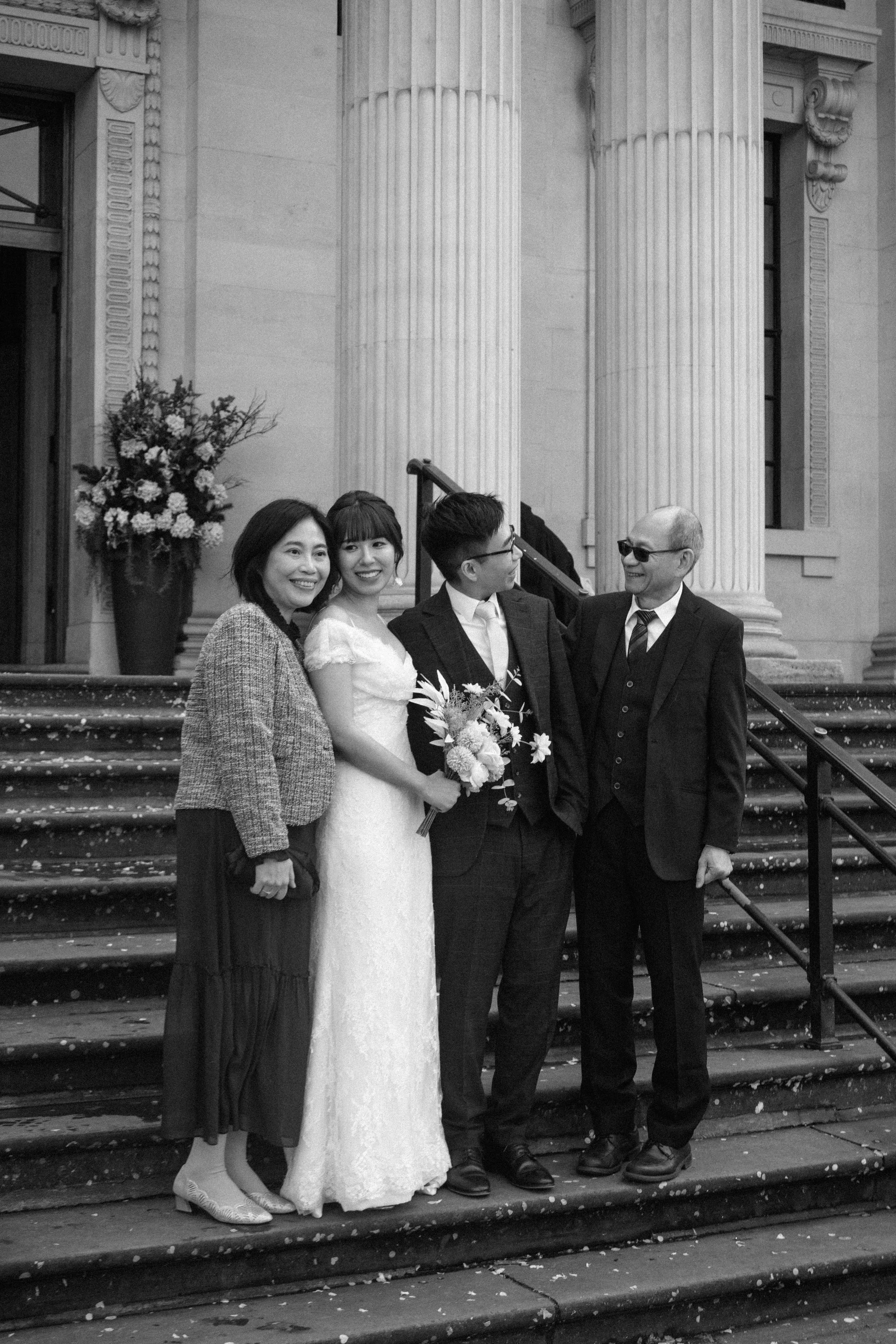 Black and white photo of a wedding group standing on stairs in front of a building with tall columns; includes a bride holding a bouquet, two women, and two men dressed in suits, smiling and looking at each other.