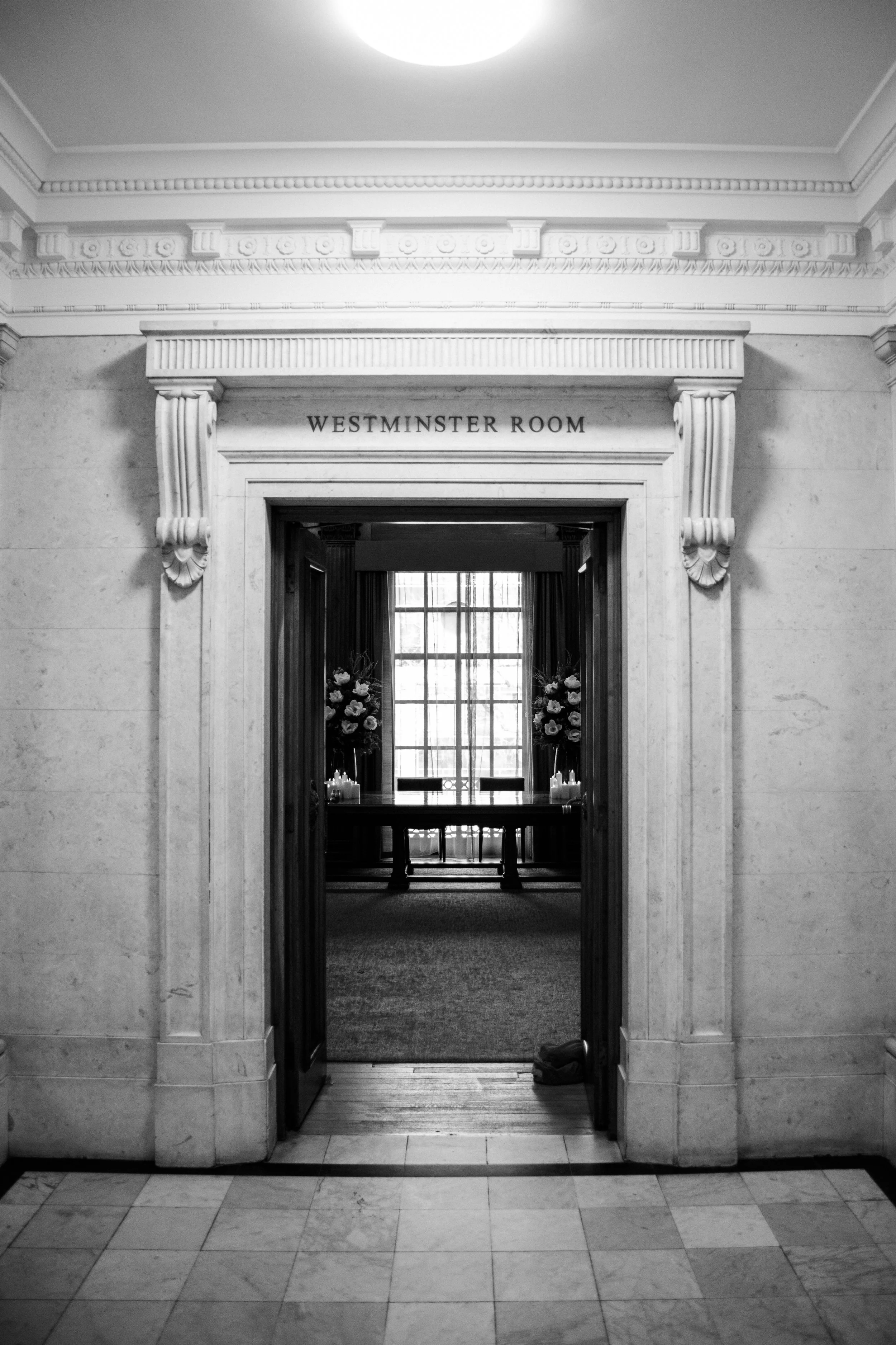 Black and white photo of an ornate doorway labeled 'Westminster Room' leading into a room with a large window, floral arrangements, and a bench.