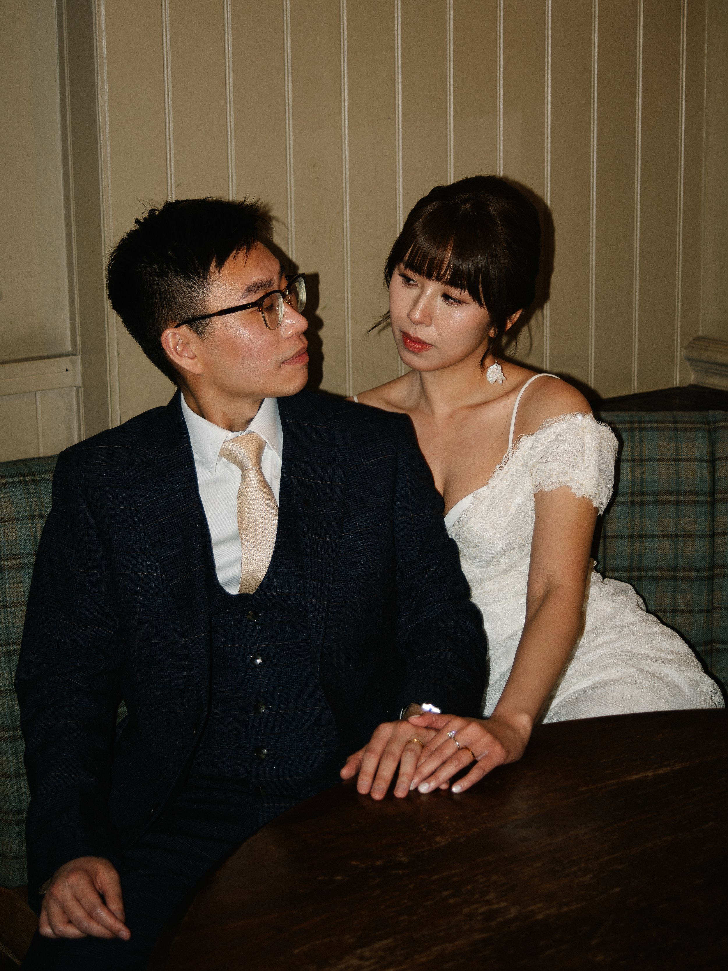 A man and woman sit close together in a cozy, dimly lit room. The woman leans toward the man, resting her hand on his. They exchange gazes with subtle affection. The man wears glasses, a dark suit, and a tie, while the woman is dressed in a white dress with lace detail and has earrings.