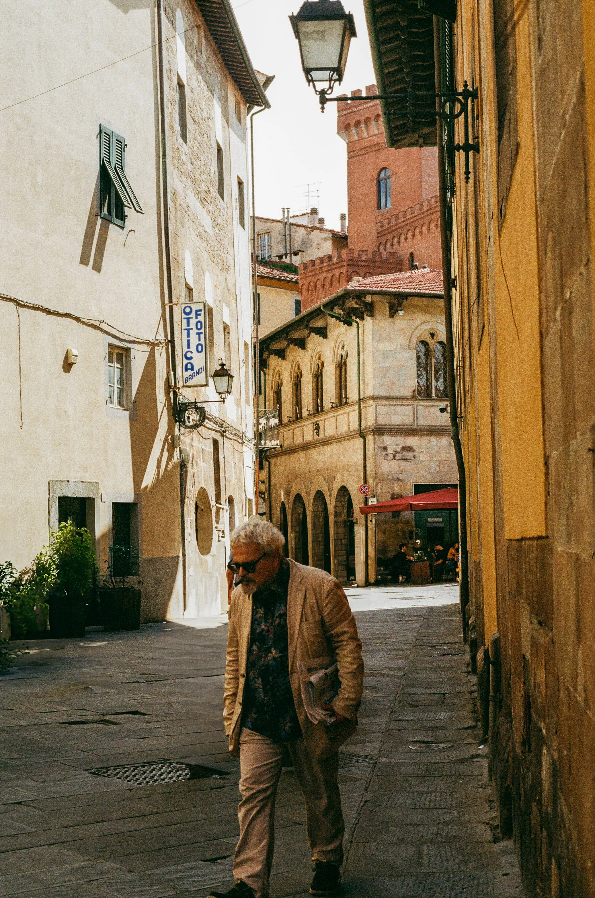 A man in a beige suit walking down a narrow street in a European city, with colorful old buildings and a tower in the background.