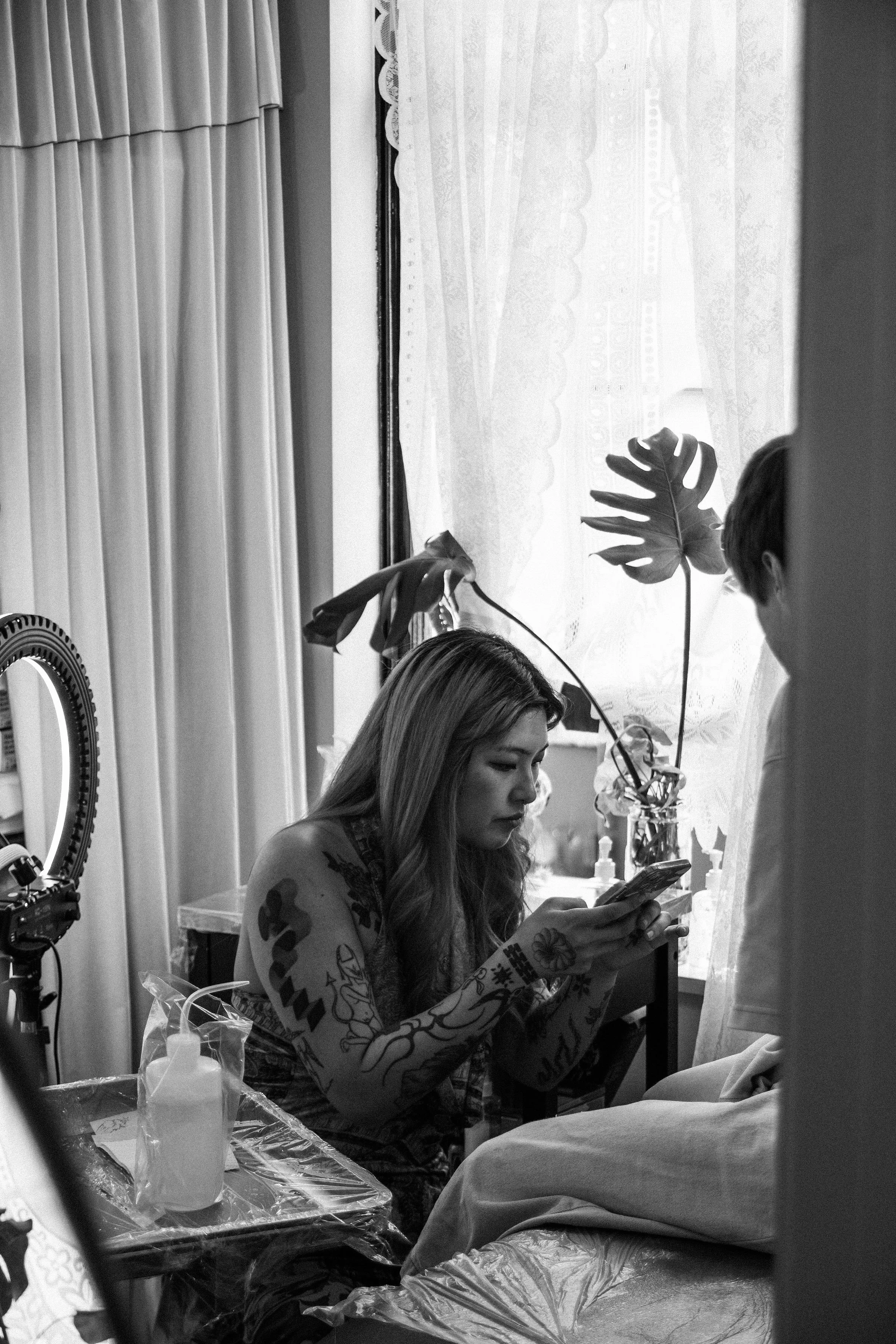 A woman with tattoos sitting in a room, looking at her phone. She has long hair and is surrounded by tattoo equipment, a large Monstera leaf, and floral curtains.