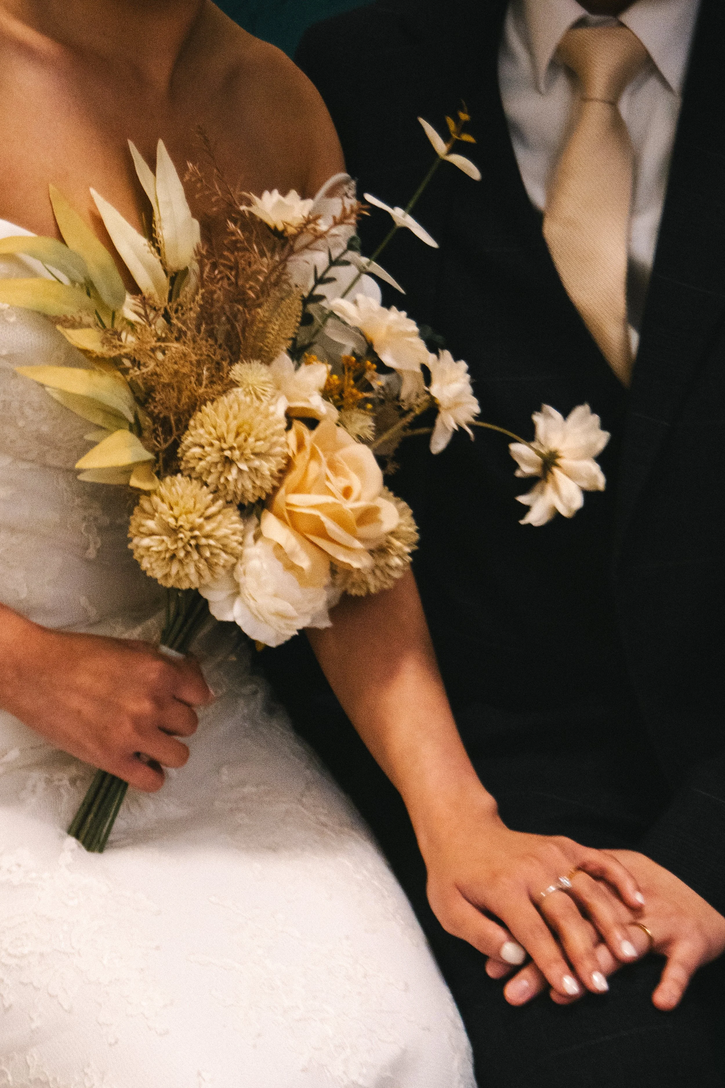 A bride and groom holding hands, with the bride holding a bouquet of cream and white flowers and the couple's wedding rings visible.