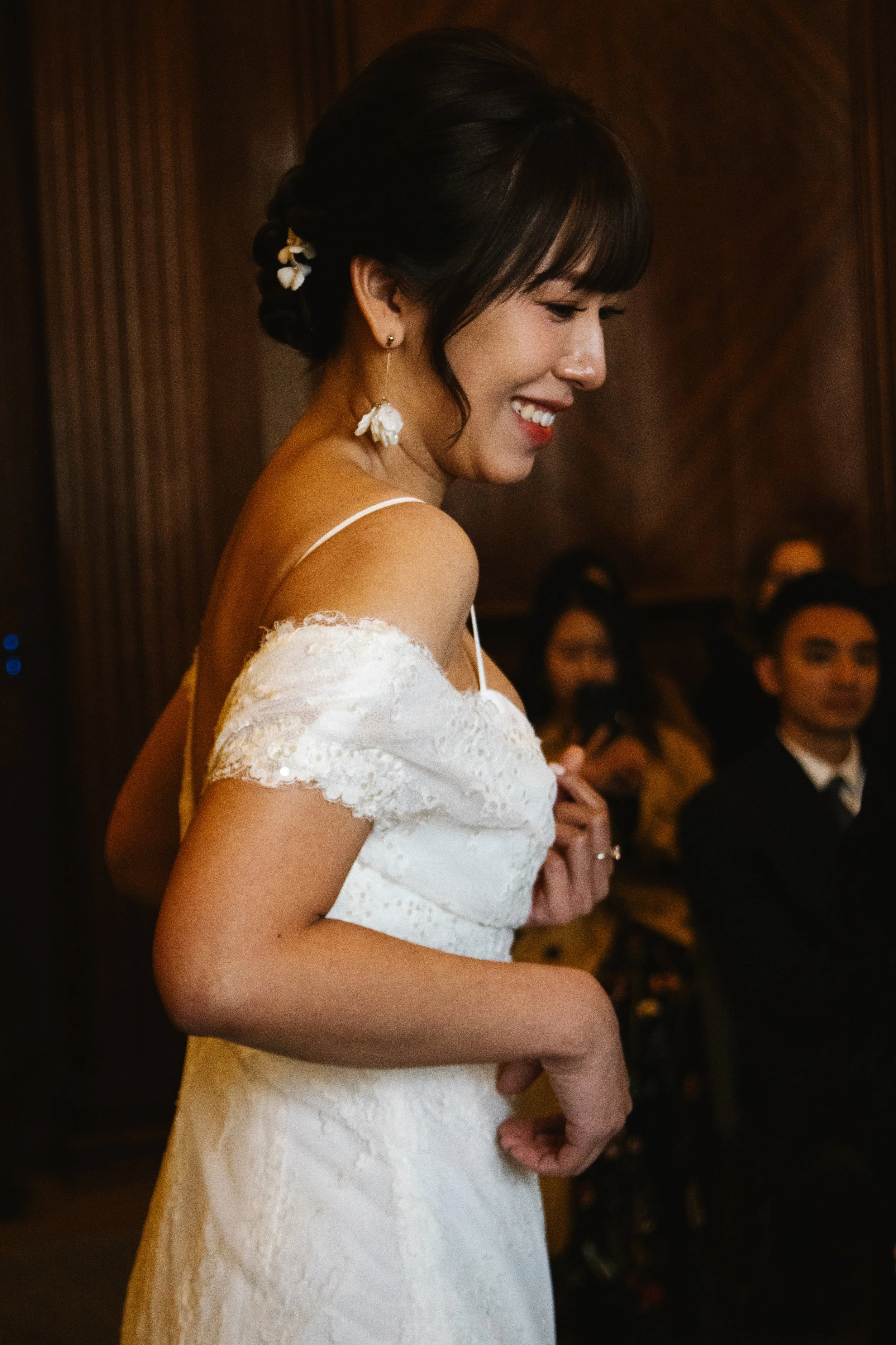 A woman in a white dress with lace details, smiling and looking down, with dark hair styled with flowers, at a formal event with guests in the background.