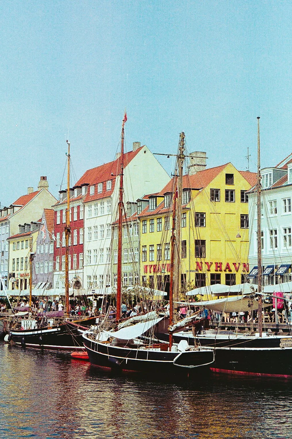 Colorful waterfront scene with sailboats docked along a pier and rainbow-colored buildings in the background under a clear sky.
