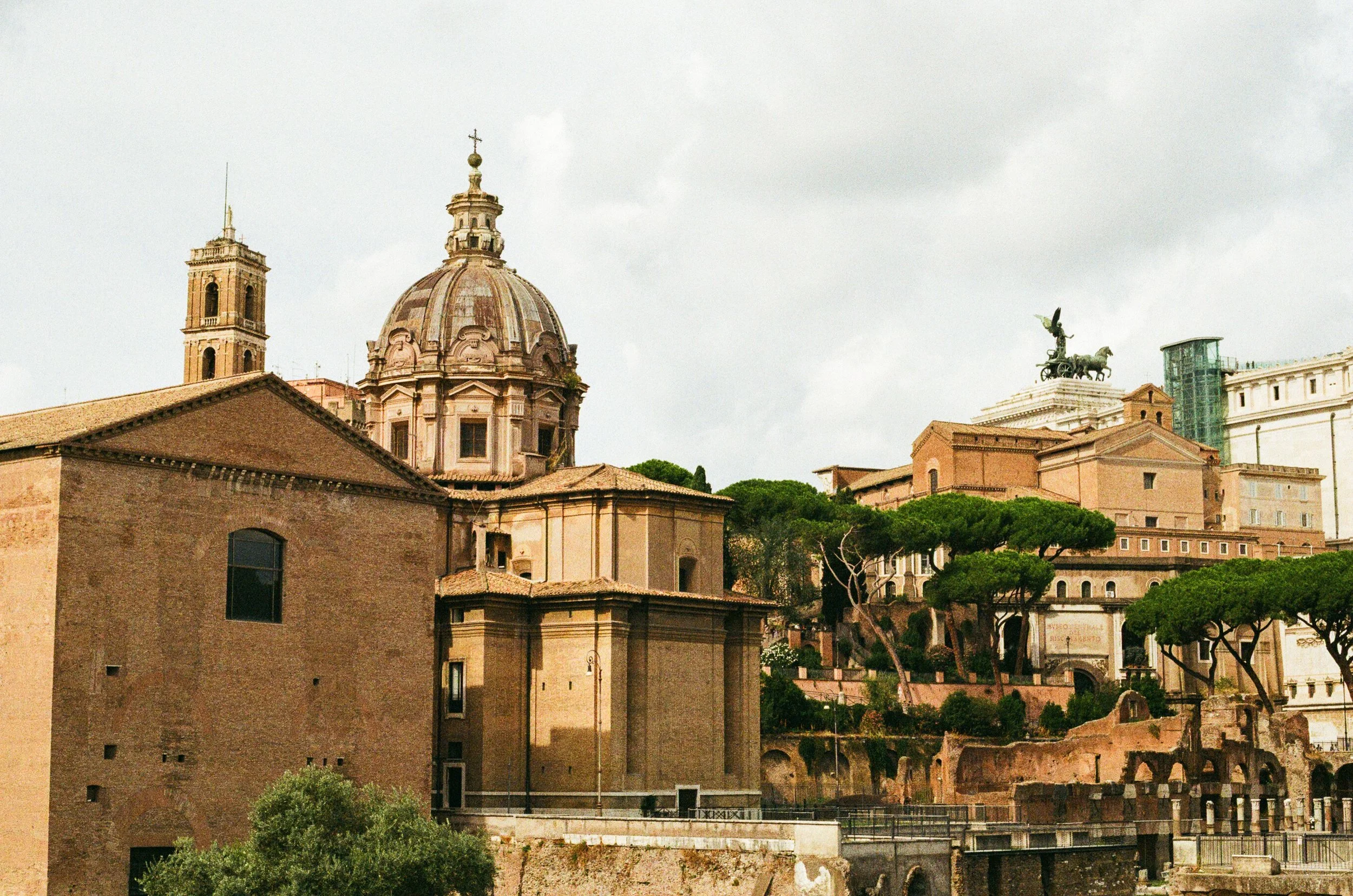 Historical cityscape with a large domed church, ancient ruins, and a statue of a chariot with winged figure on top, surrounded by buildings and trees.