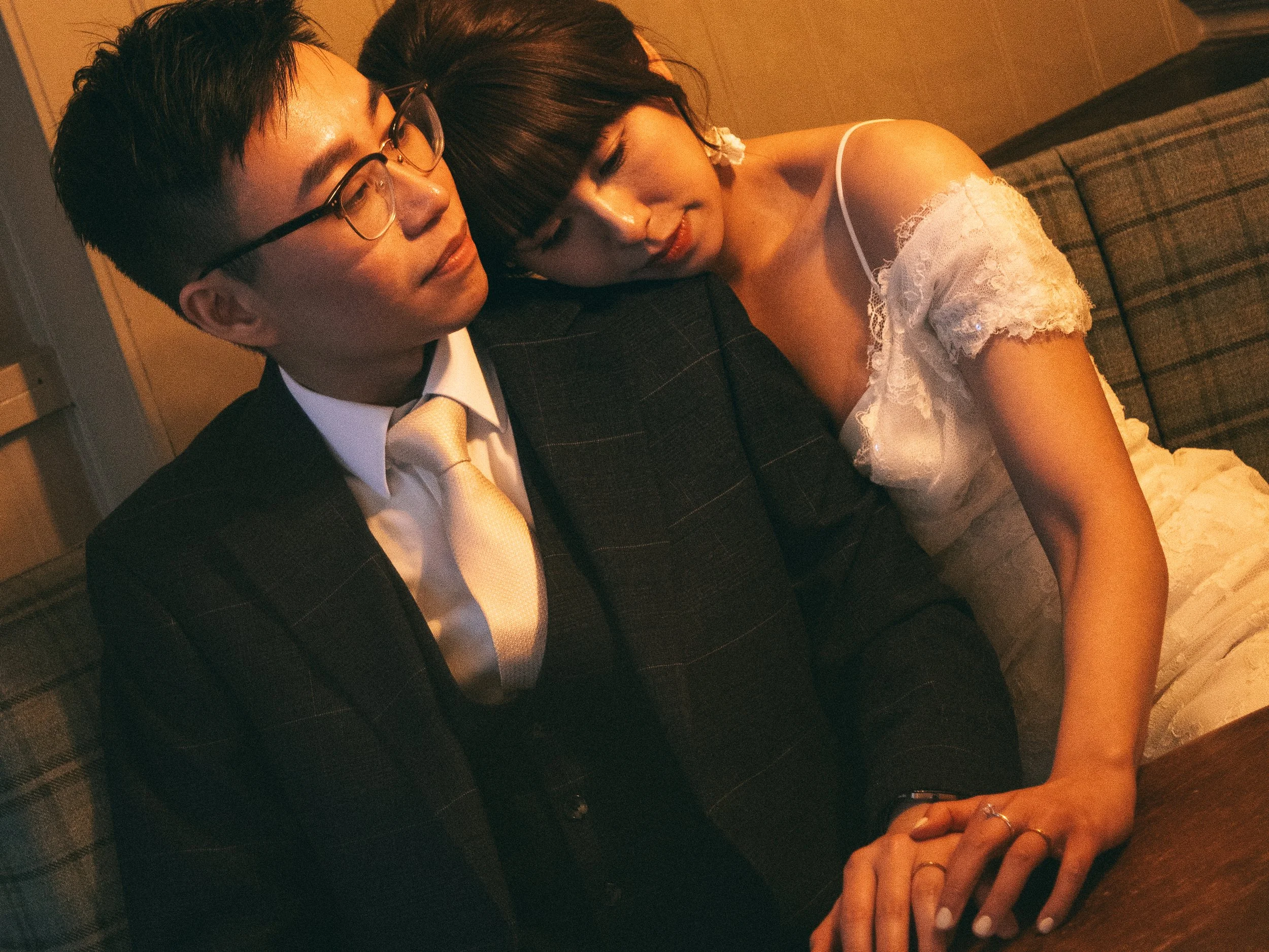 A man and woman sitting closely together on a couch, with the woman resting her head on the man's shoulder. The man is wearing glasses, a dark suit, a white shirt, and a light-colored tie. The woman is wearing a white or cream dress with lace details and has her eyes closed.