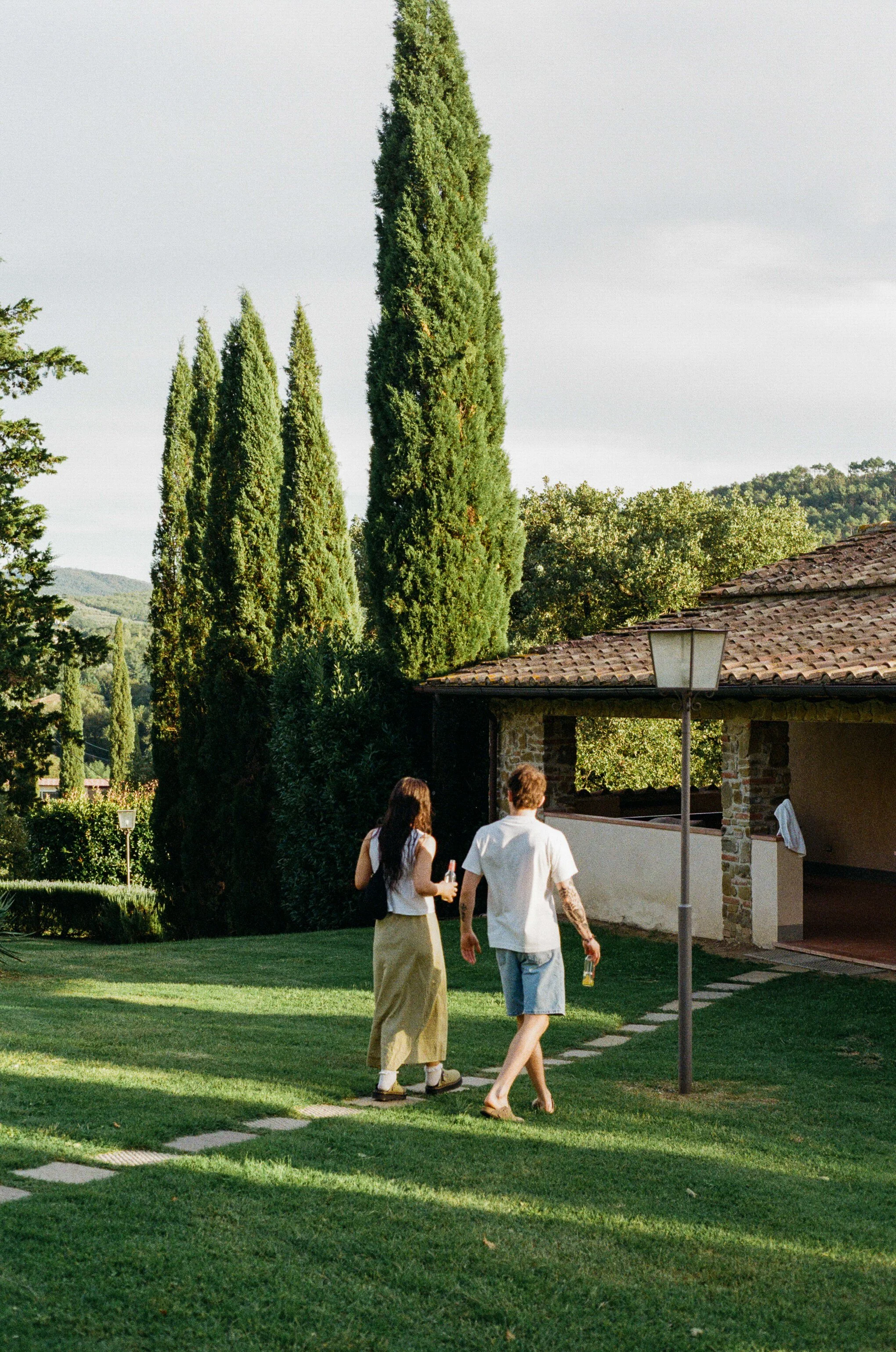 Two people are walking on a stone pathway in a lush green yard with tall trees and a stone building with an outdoor overhang. One is a woman with long dark hair, wearing a white top and beige skirt, holding a drink, and the other is a man with short hair, wearing a white shirt and denim shorts, also holding a drink.