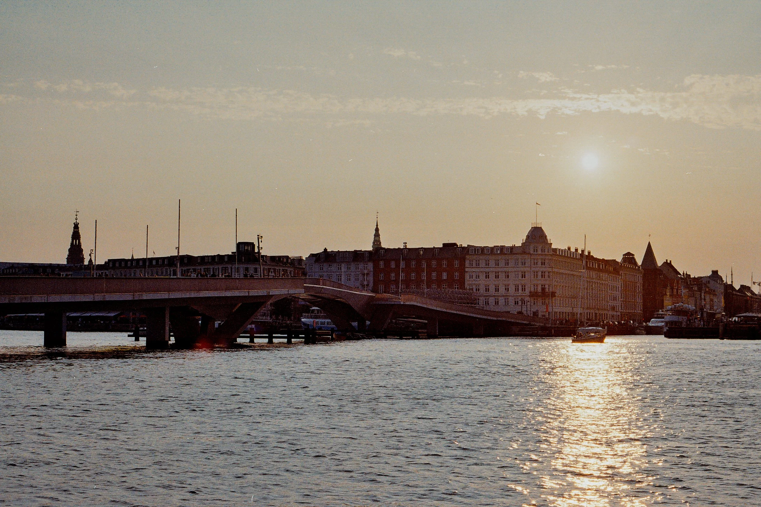 Sunset over a city river with buildings and a bridge in the background.