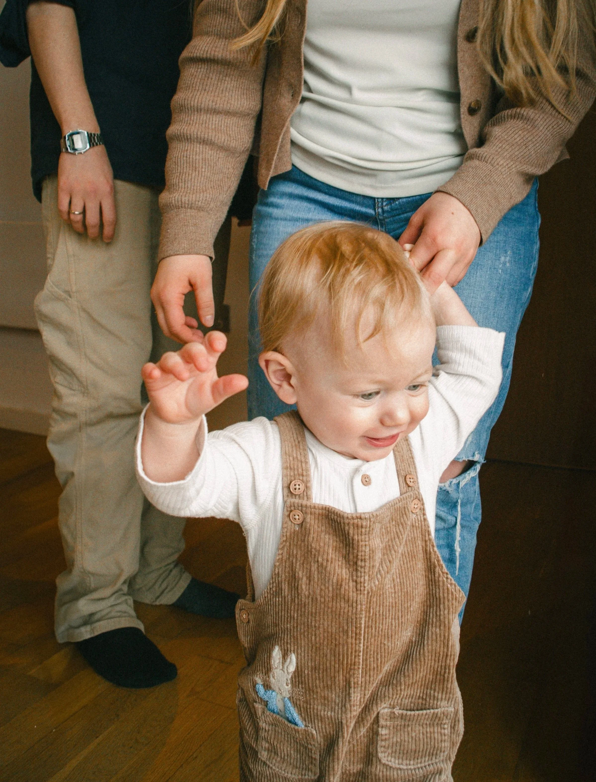 A young child with red hair wearing brown overalls and a white shirt stands and raises one arm as an adult gently holds their hand. Another person stands nearby in beige pants and a black shirt, with a hand resting on their hip.