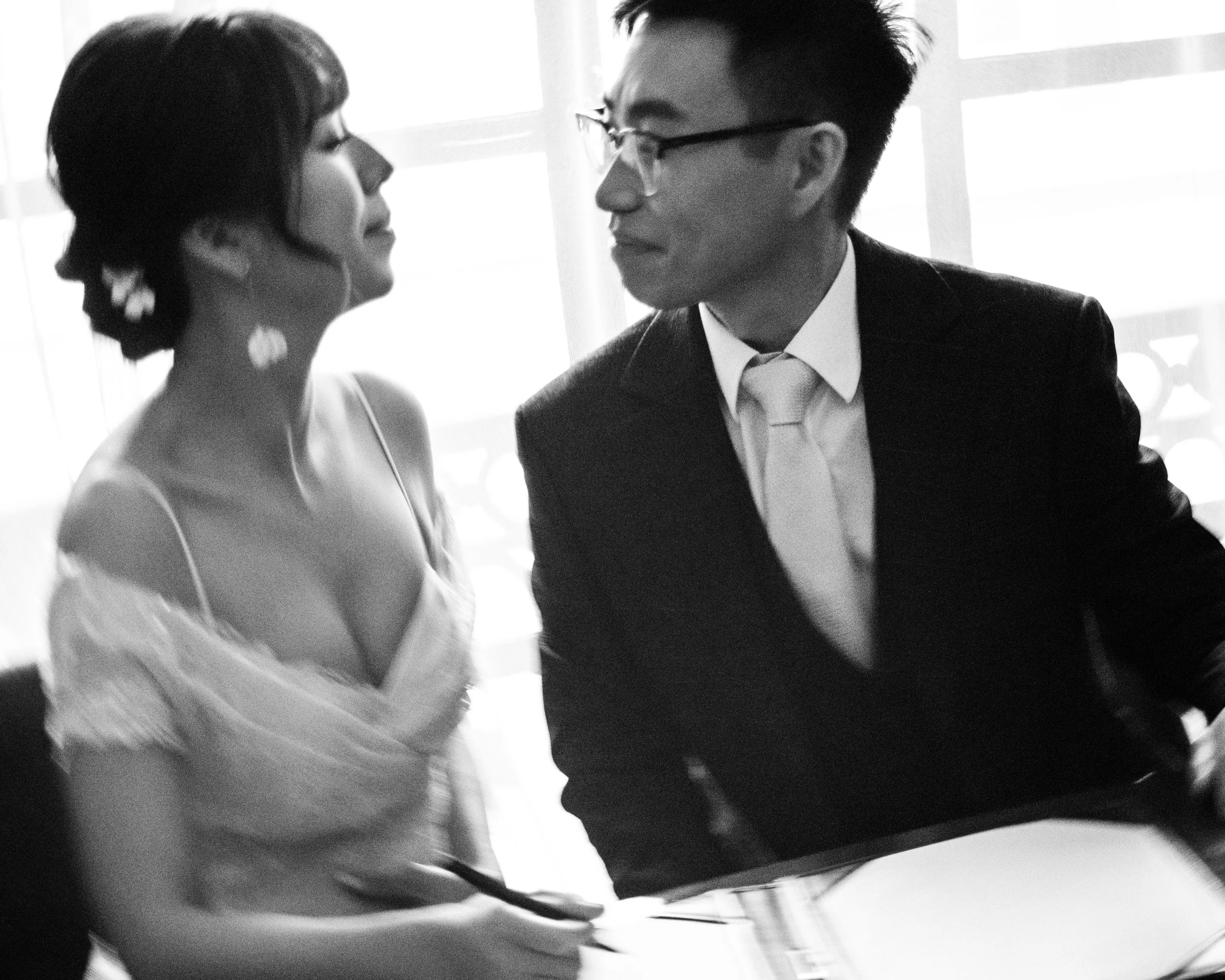 A black and white photo of a woman and a man sitting close, looking at each other. The woman wears an off-shoulder dress, and the man wears a suit with a tie. They appear to be engaged in an intimate or romantic moment.