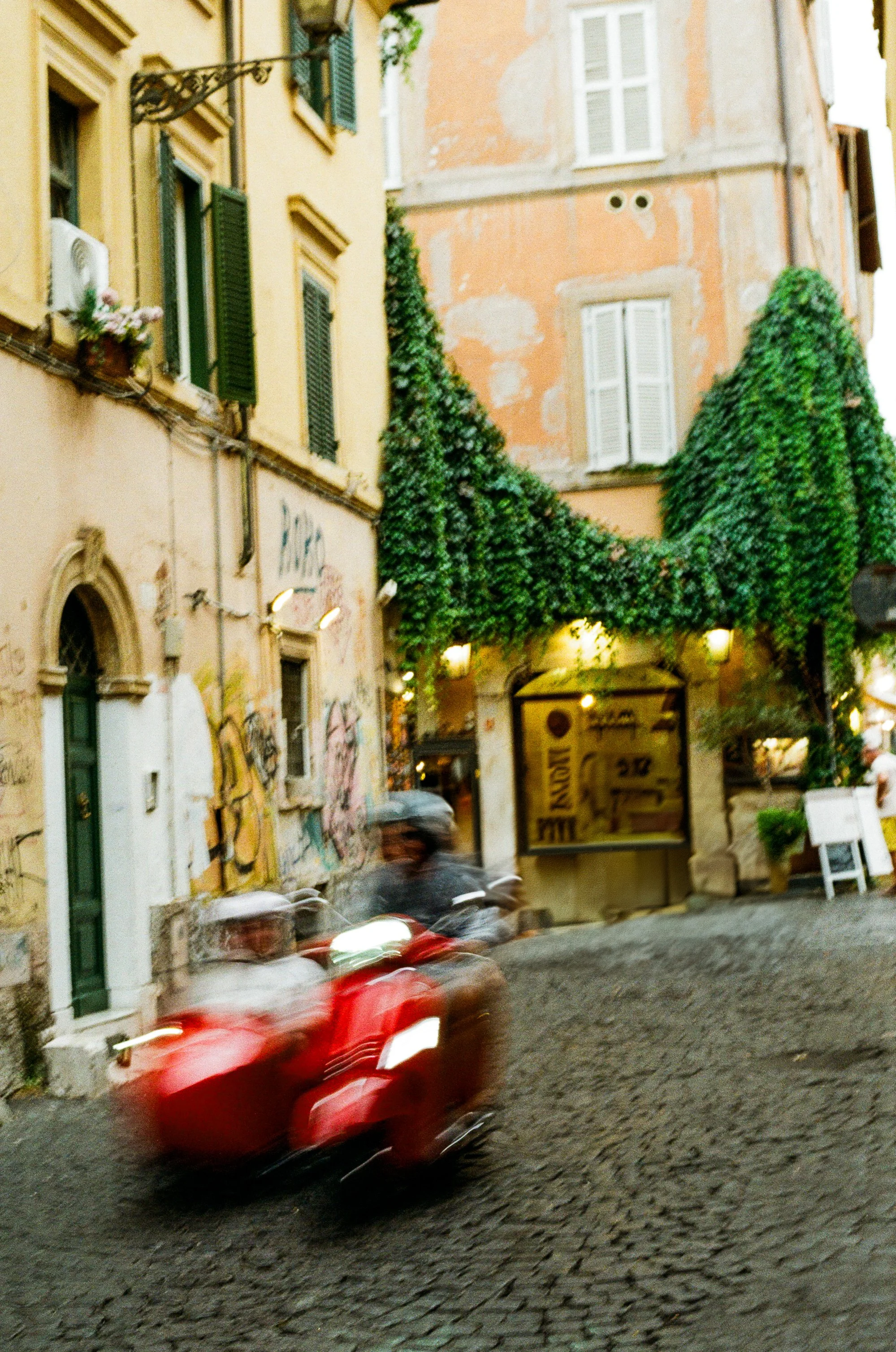 A blurry photo of two people riding a red Vespa scooter on a cobblestone street in a European city. The scene includes colorful old buildings with green shutters and decorated facades, and a building covered in green ivy.