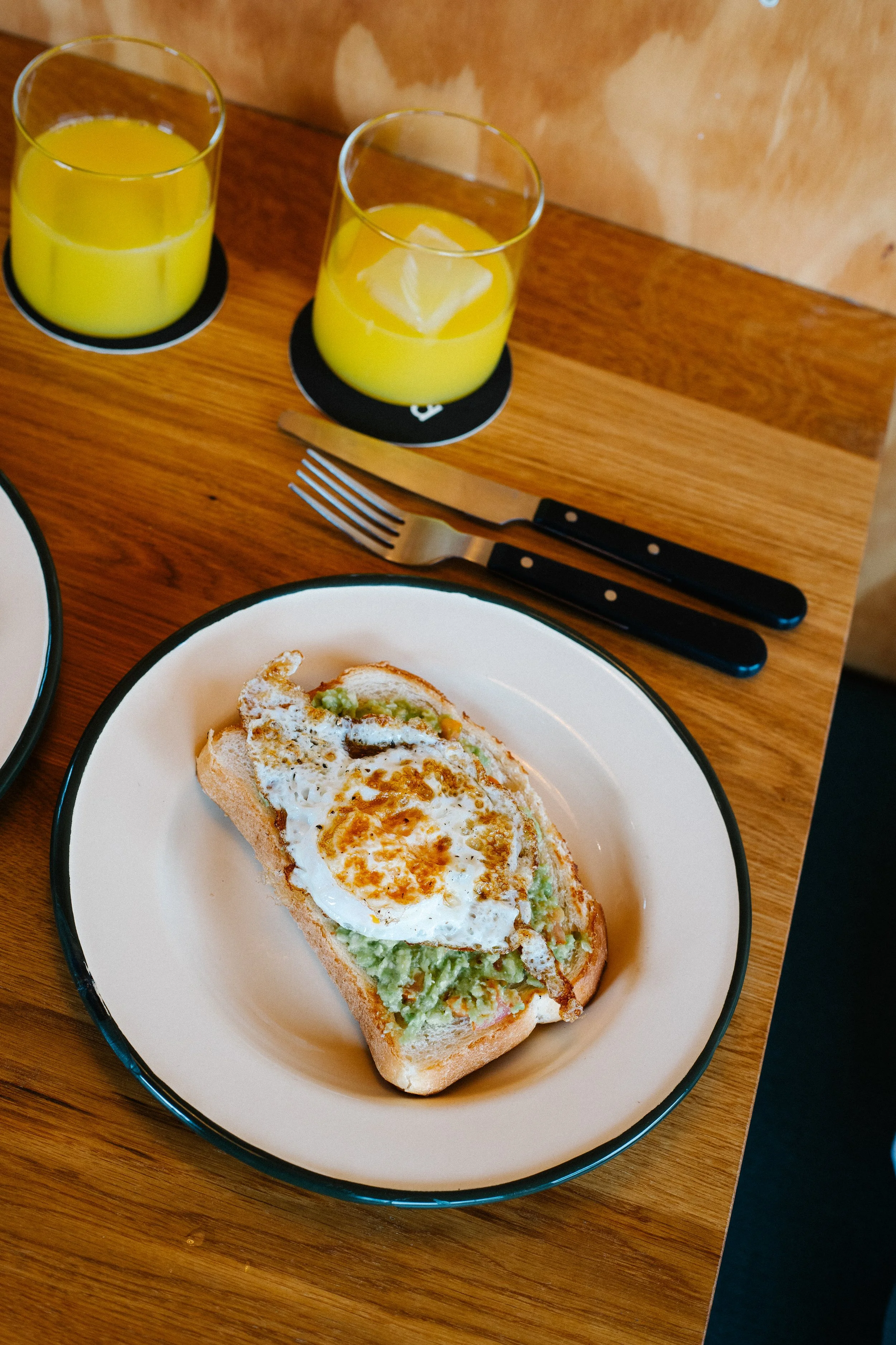 A plate with avocado toast topped with a fried egg, two glasses of orange juice with ice on black coasters, and a fork and knife on a wooden table.