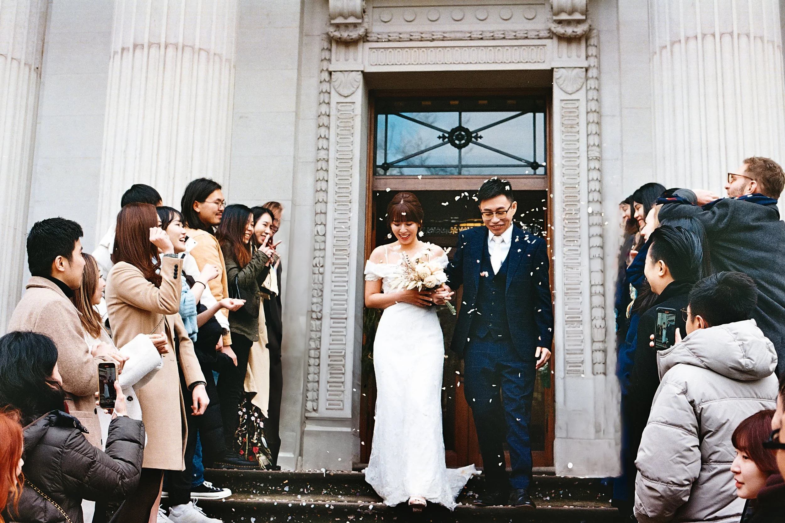 A bride and groom walking out of a building surrounded by friends and family, celebrating their wedding, with confetti falling around them.