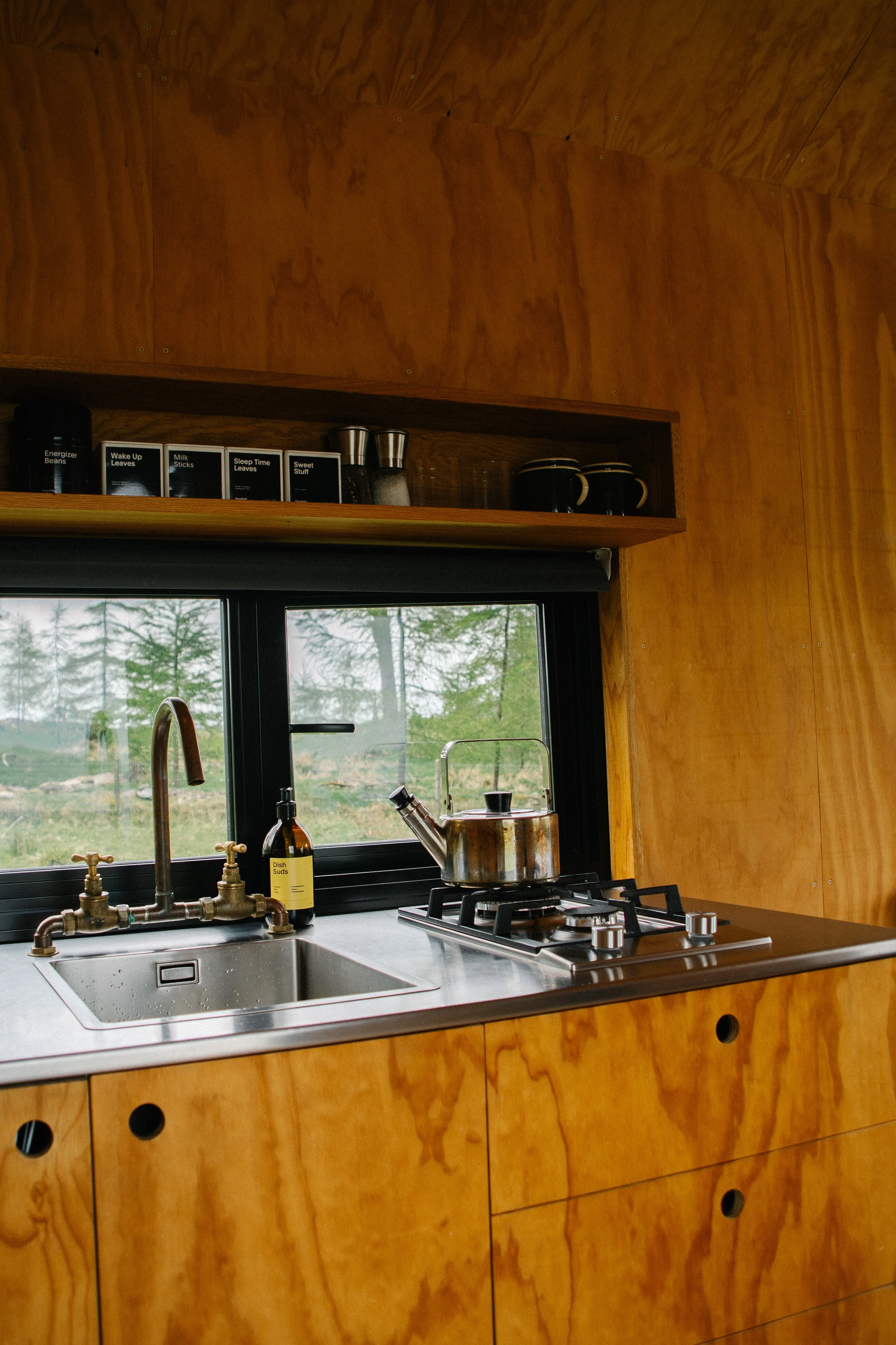 Kitchen with wooden cabinets, stainless steel sink, stove with pot, and window showing trees outside.