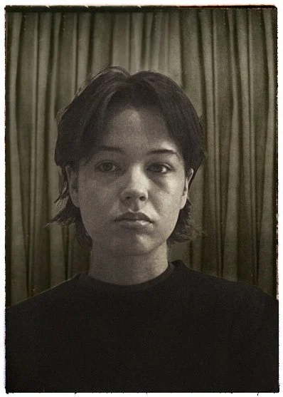 Black and white portrait of a person with short dark hair, wearing a black shirt, standing in front of a curtain.