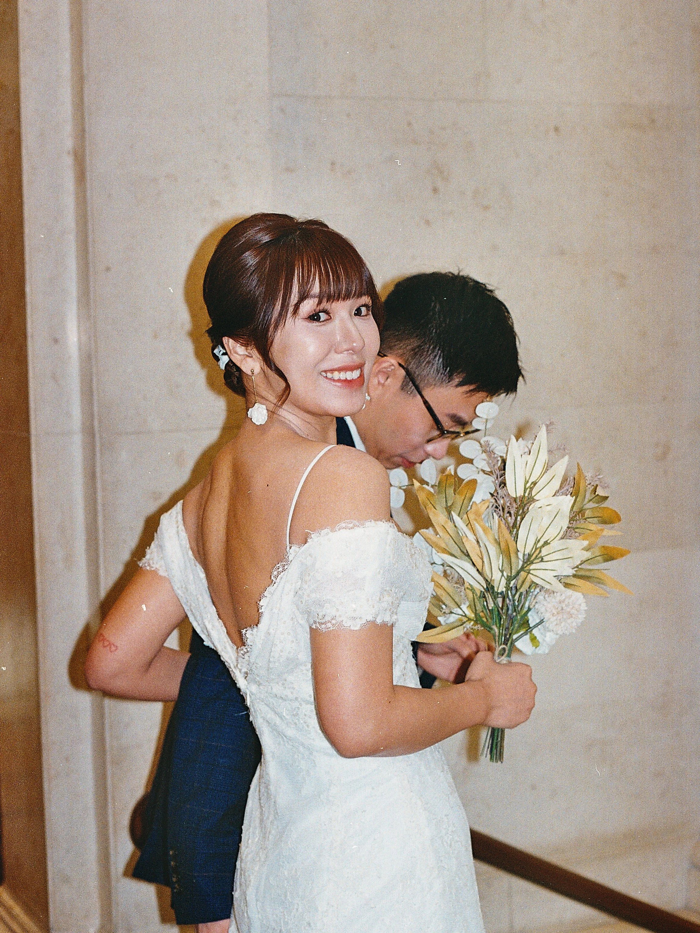 Woman in a white lace wedding dress holding a bouquet of flowers, smiling, with a man in a suit and glasses behind her, in an indoor setting.