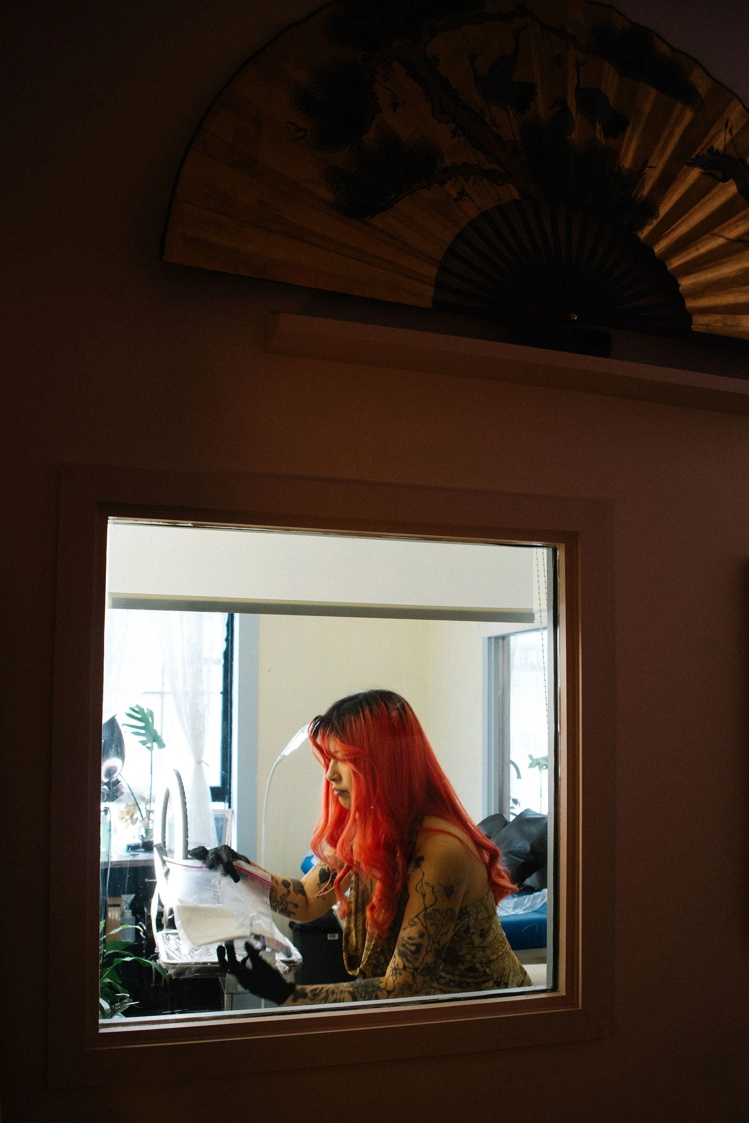 A woman with red hair and tattoos, wearing black gloves, sitting near a window inside a room, looking at a paper or magazine.