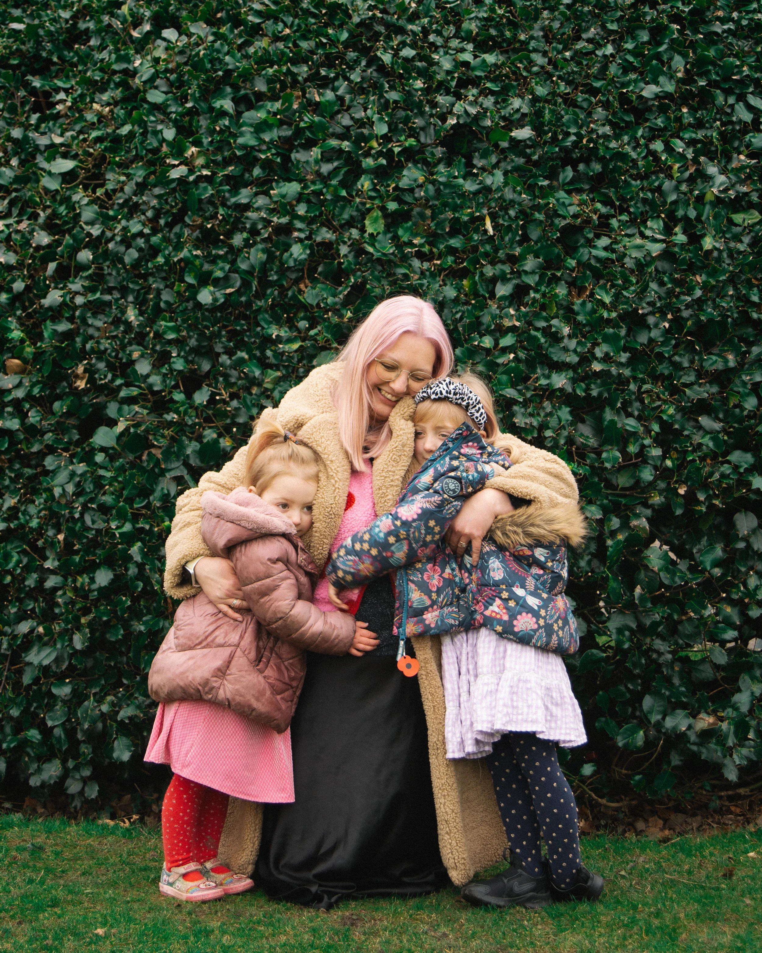 A woman with pink hair and glasses hugging three young girls in front of a leafy hedge, all dressed in warm clothing and smiling.