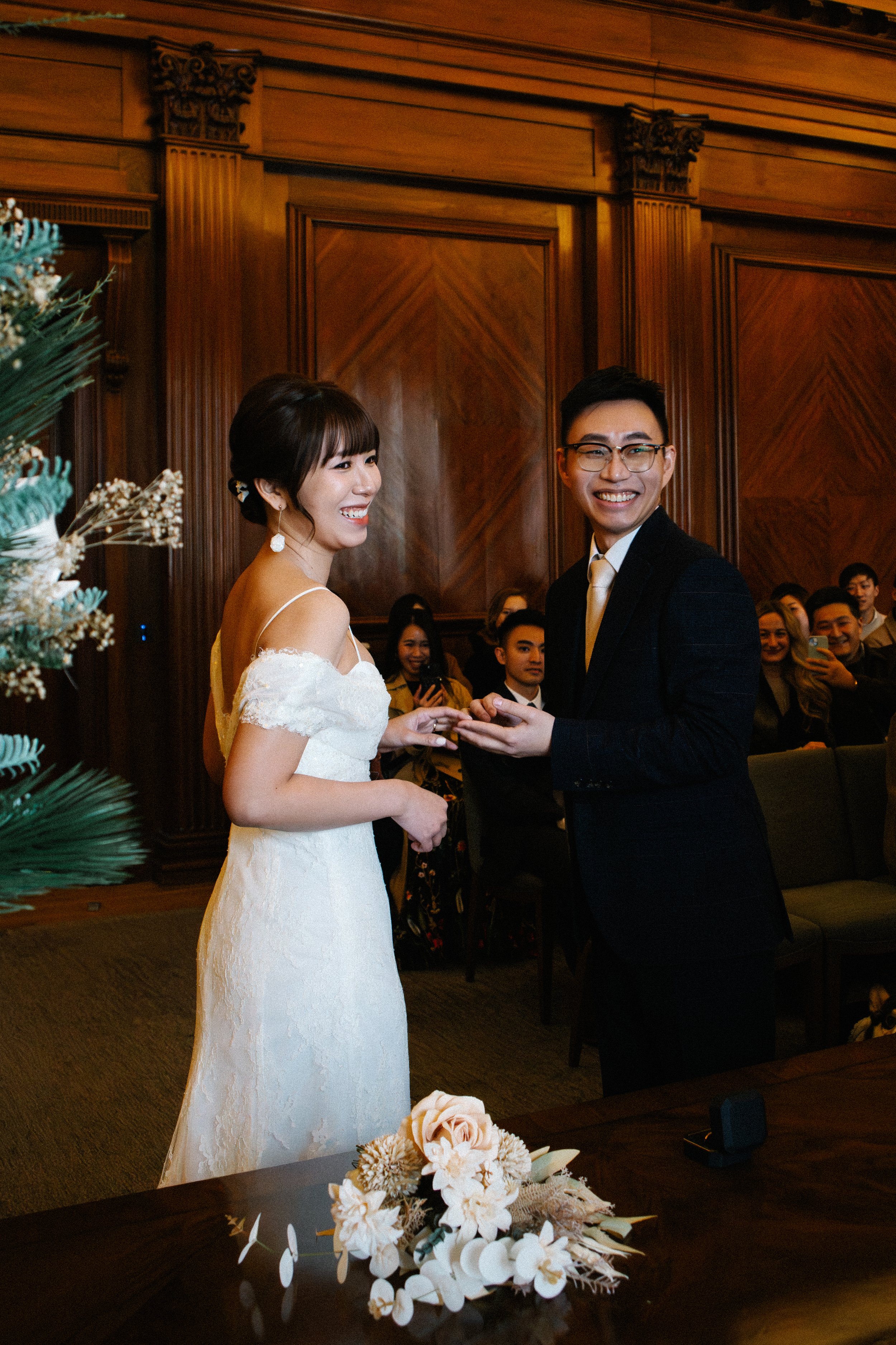 A bride and groom exchanging rings at their wedding ceremony, with guests seated behind them in a wood-paneled room, and a bouquet of flowers on the table in front.