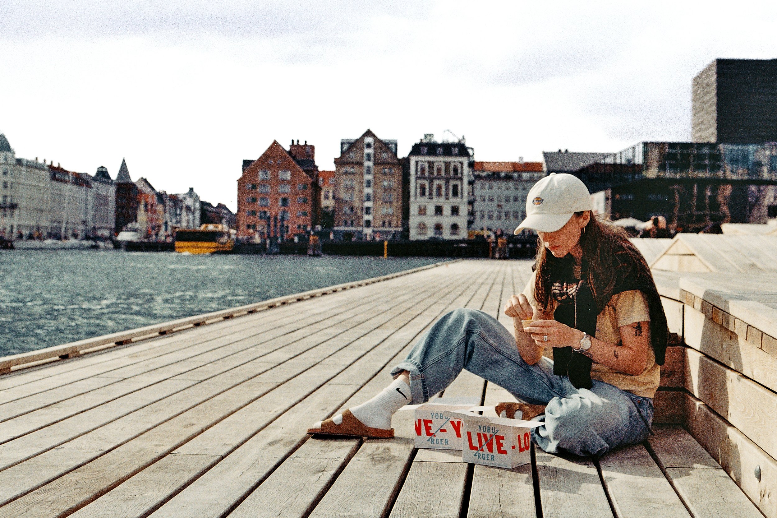 A woman with brown hair, wearing a beige cap, black vest, beige t-shirt, blue jeans, and white socks with Nike sandals, sitting on a wooden dock by the water, looking at her phone. A pizza box with red and blue text is next to her.
