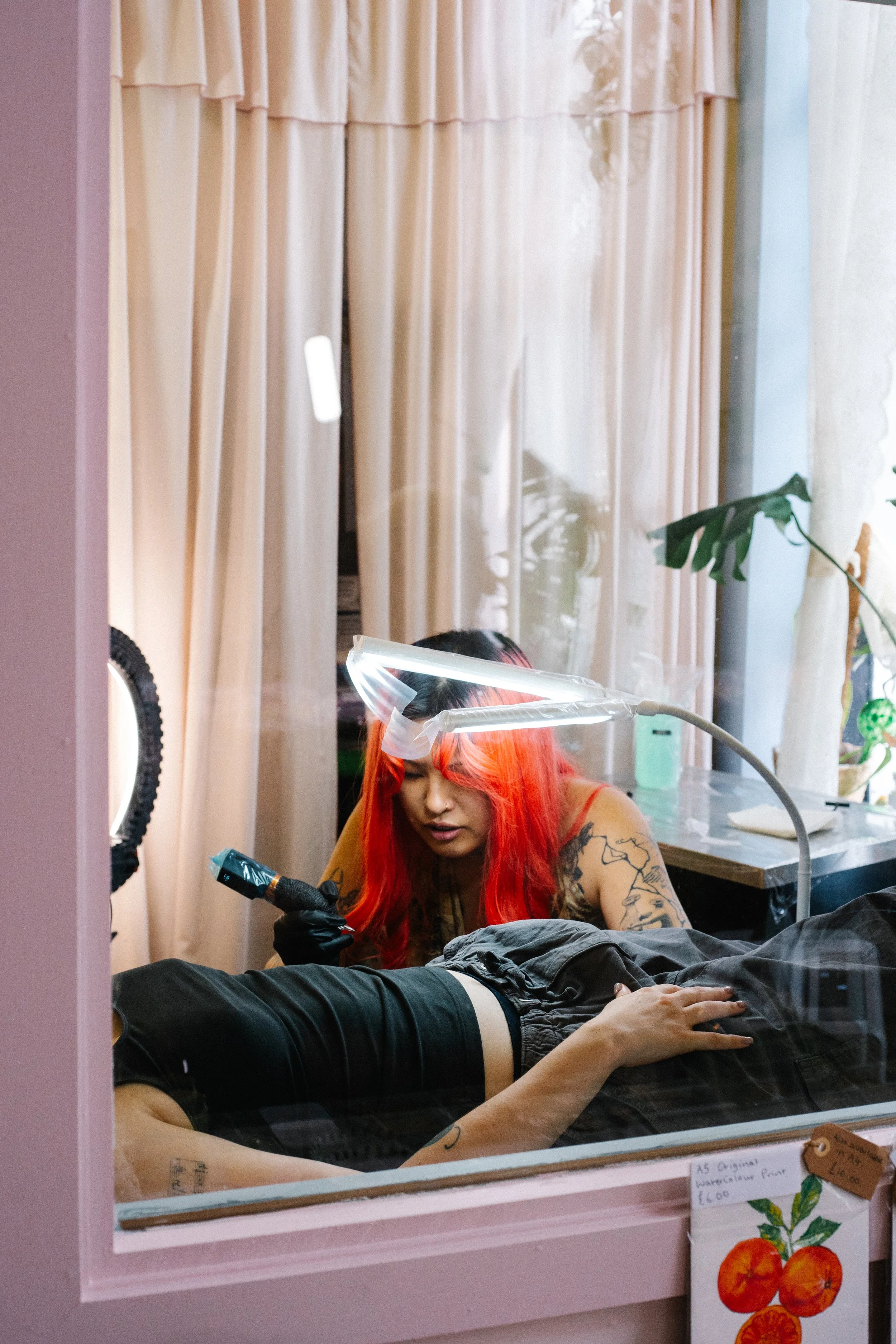 Tattoo artist with long red hair working on a customer's arm, working inside a tattoo studio with pink walls and beige curtains.