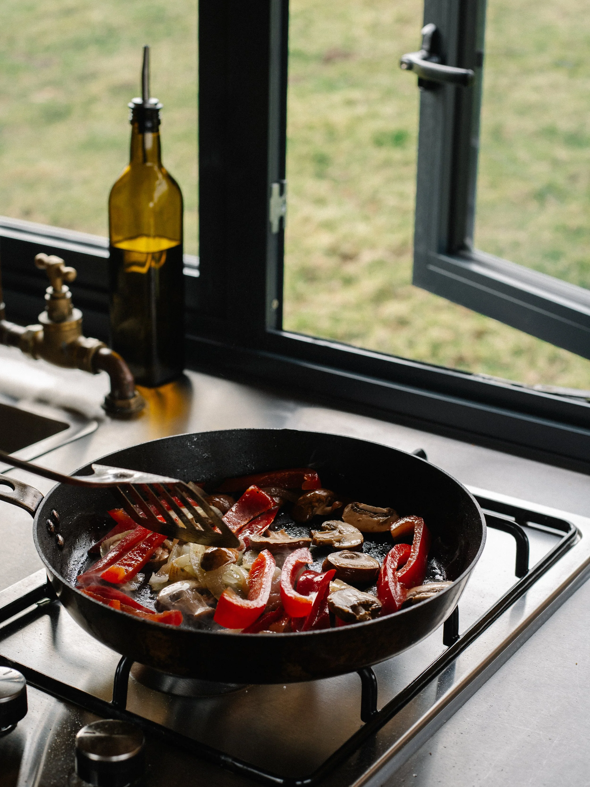 A skillet on a stove with sliced red bell peppers, onions, and mushrooms cooking. A window with an open sash is in the background.